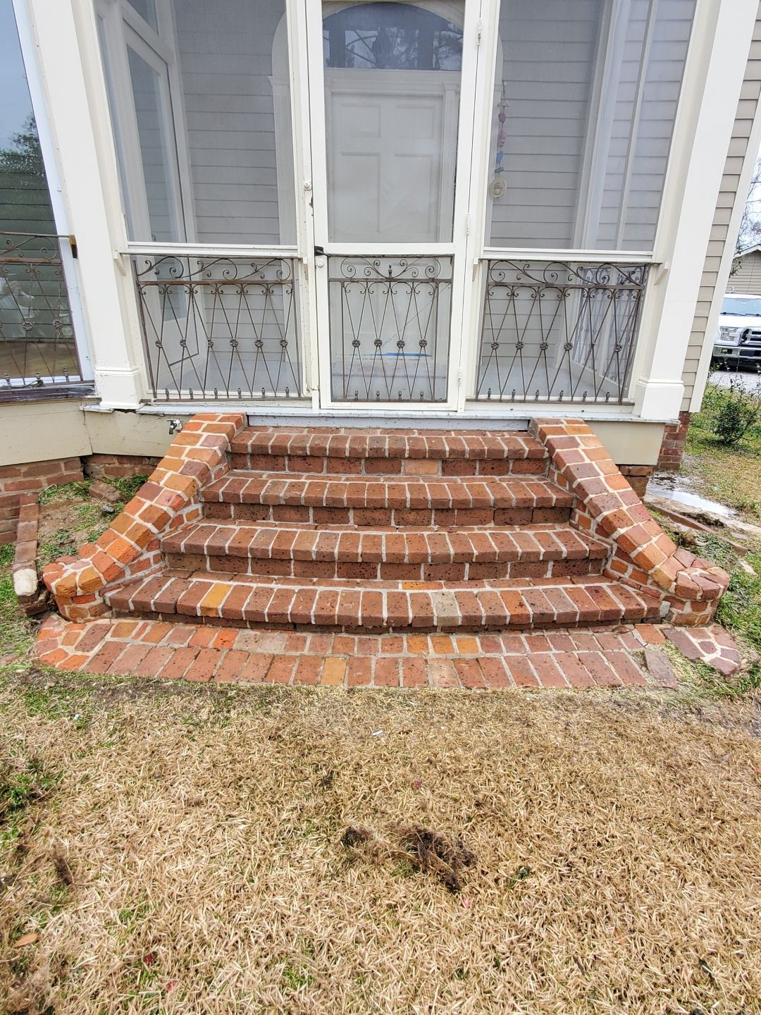 A set of three reddish-brown brick steps leading up to a screened-in porch entrance on a house exterior.