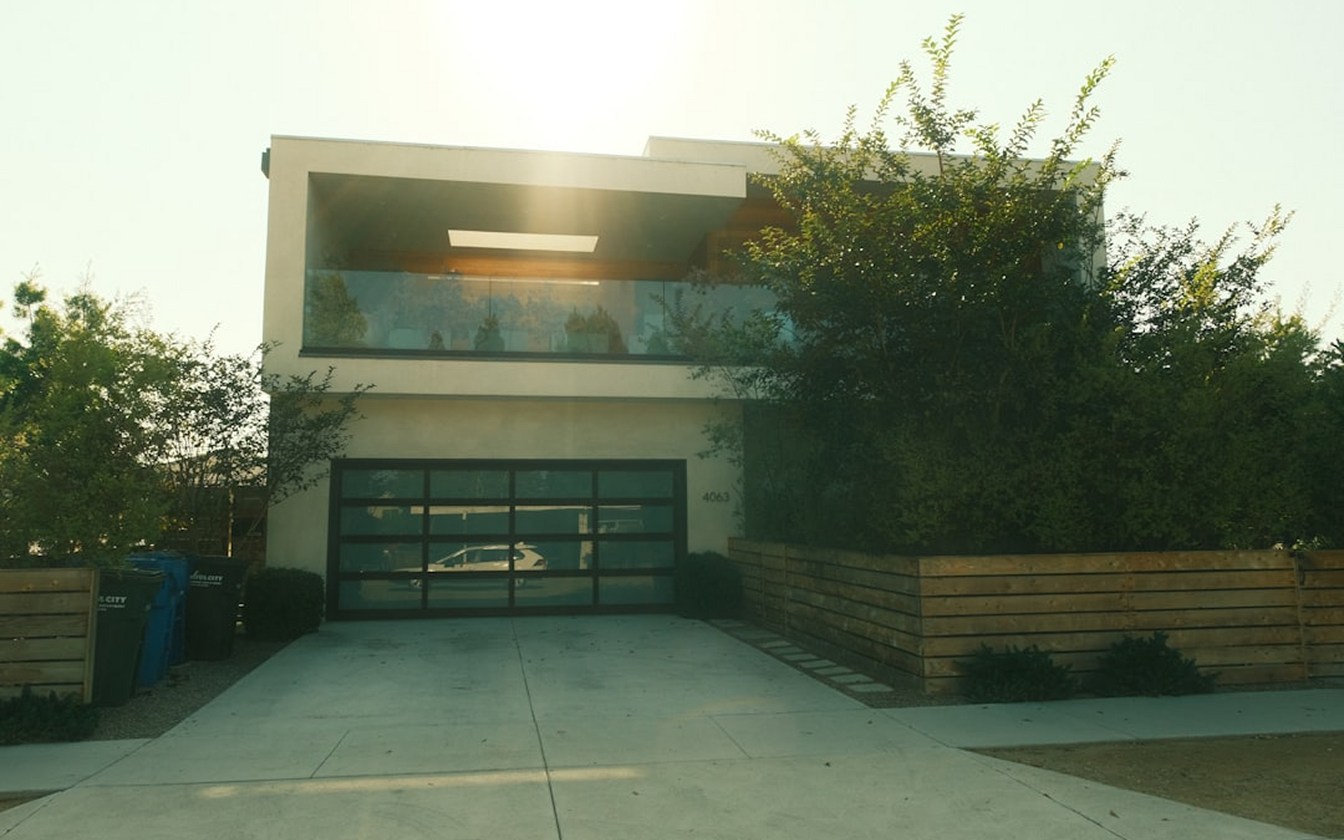 Modern two-story house with a flat roof, large glass garage door, and a horizontal wood fence under bright sunlight.