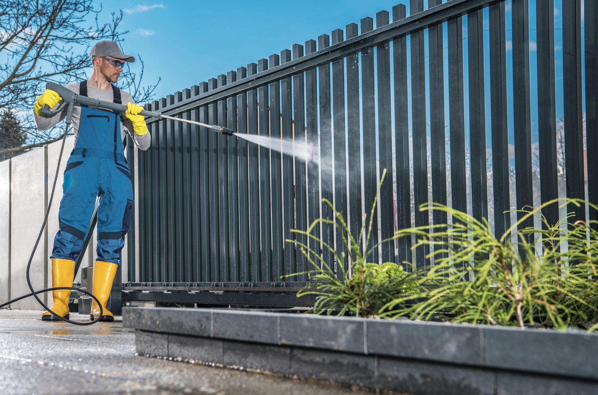 A person wearing blue overalls and yellow boots uses a pressure washer to clean a dark metal fence outdoors.