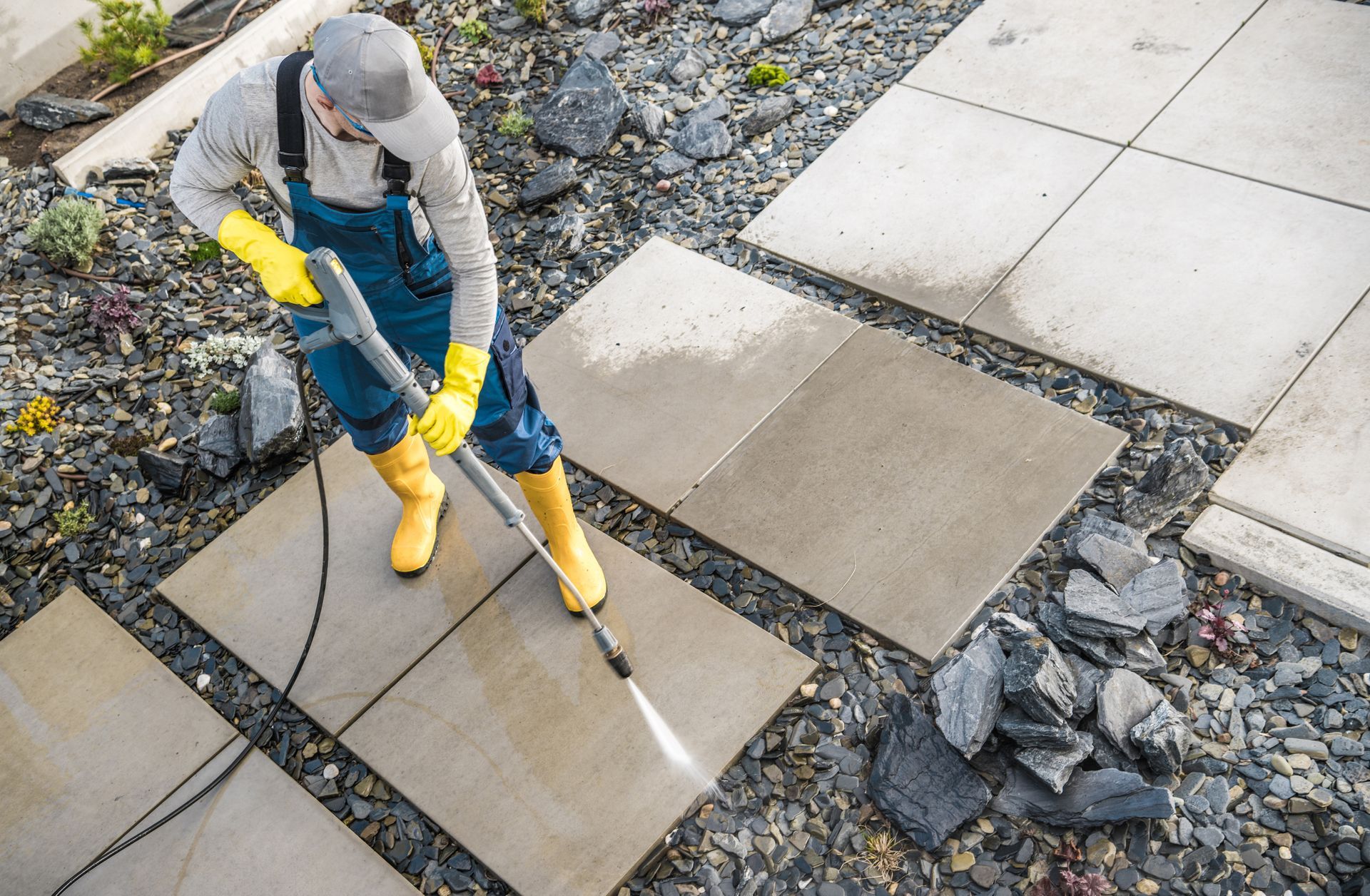 A worker in blue overalls and yellow boots uses a power washer to clean light-colored stone pavers in a garden.