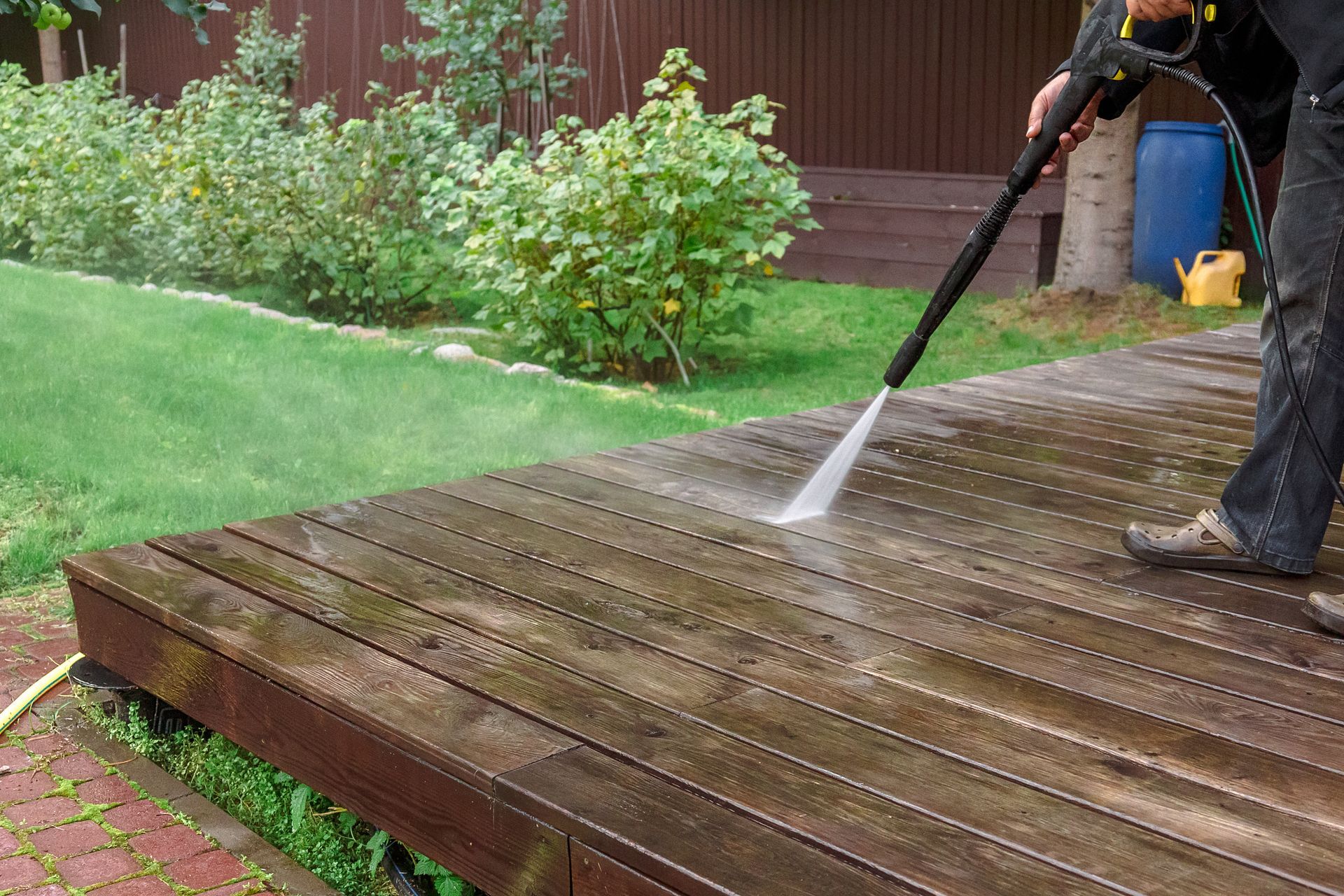 A person pressure washing a weathered wooden deck in a backyard with green grass and shrubs in the background.
