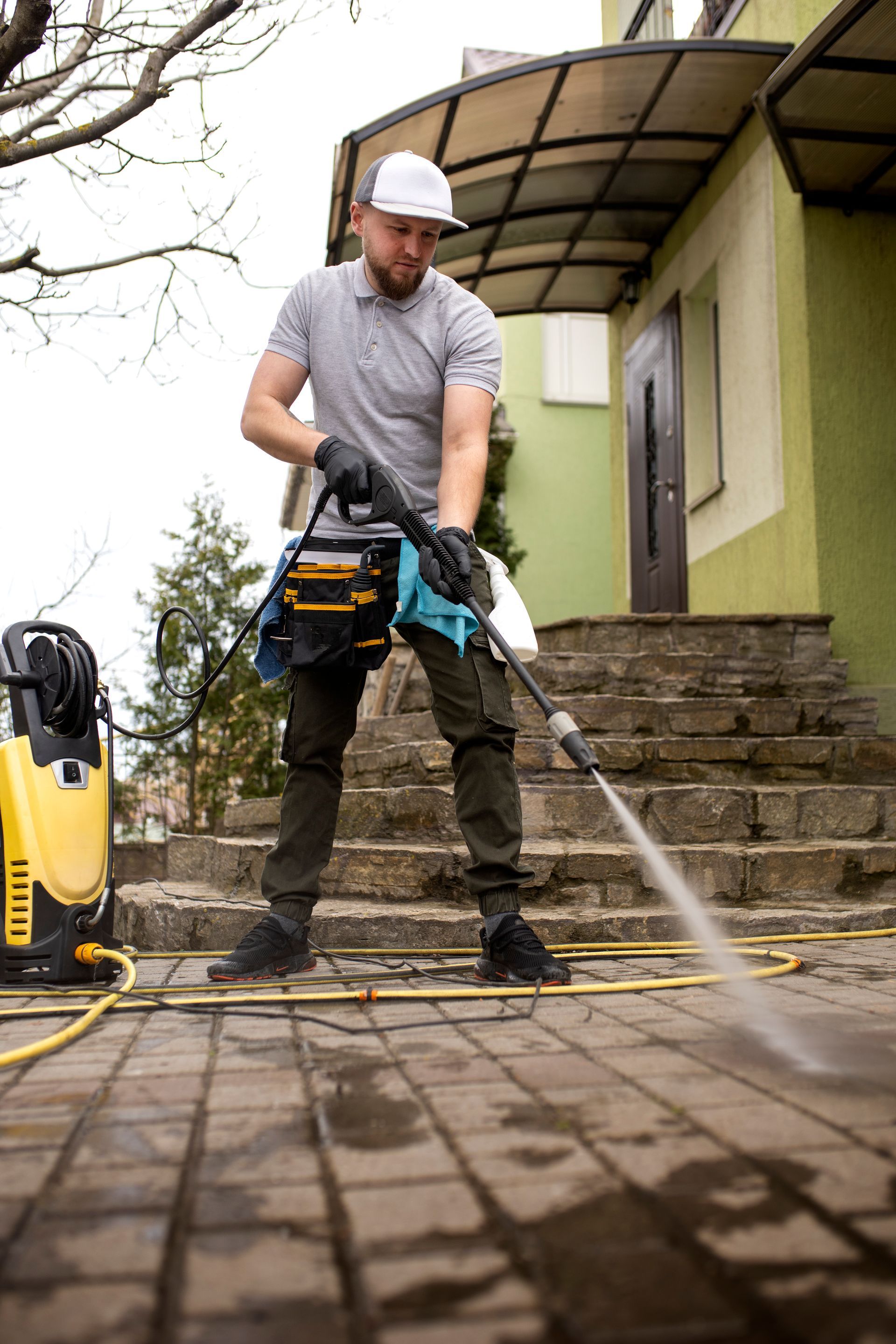 A person in a gray shirt and cap uses a yellow power washer to clean a brick walkway in front of a house.