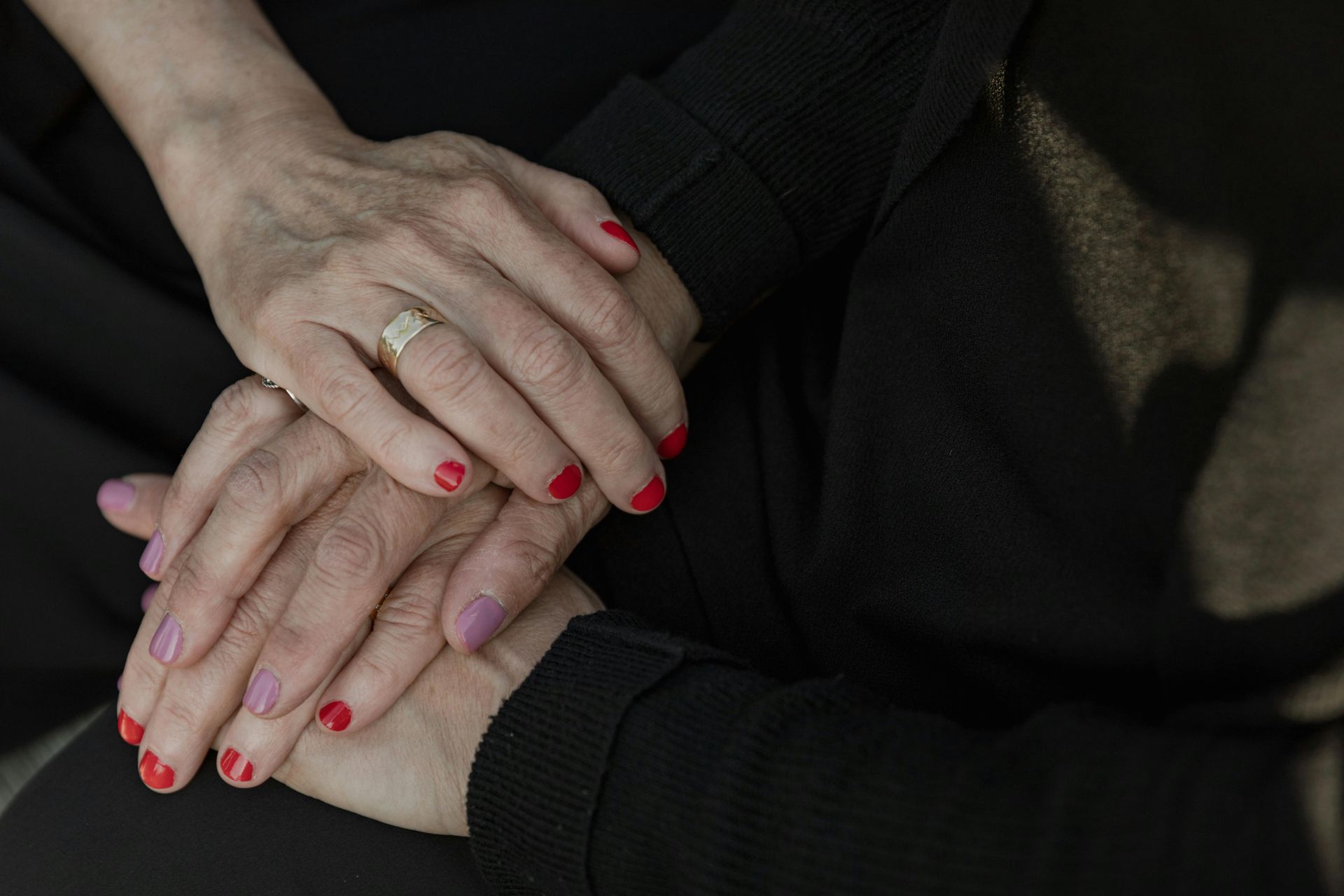 Two hands stacked together, one with red nails and a wedding ring, on a dark background