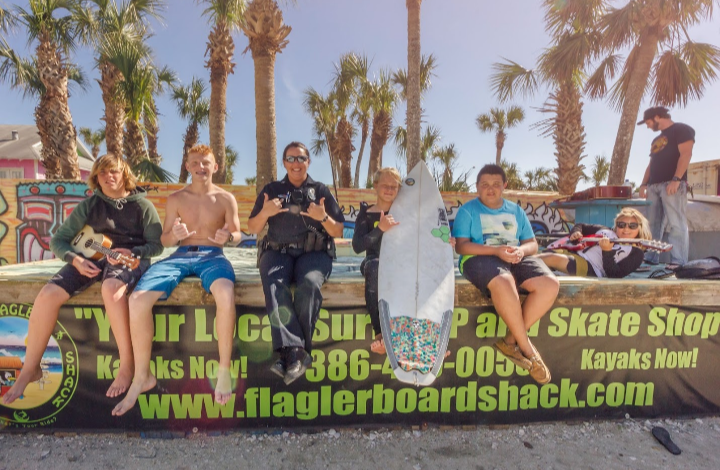 Kids sitting on skateboards with a skakeboard shop banner at an outdoor event with palm trees.