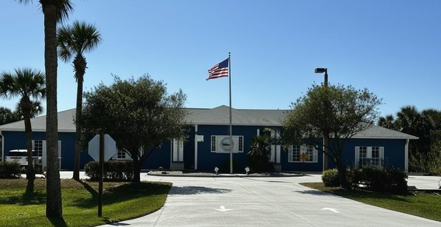 Blue building with an American flag and palm trees in front, viewed from a driveway under clear skies