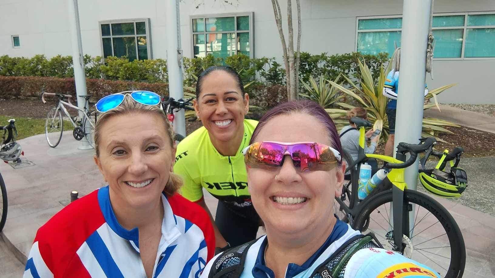 Three smiling cyclists posing with bicycles outdoors in front of a building
