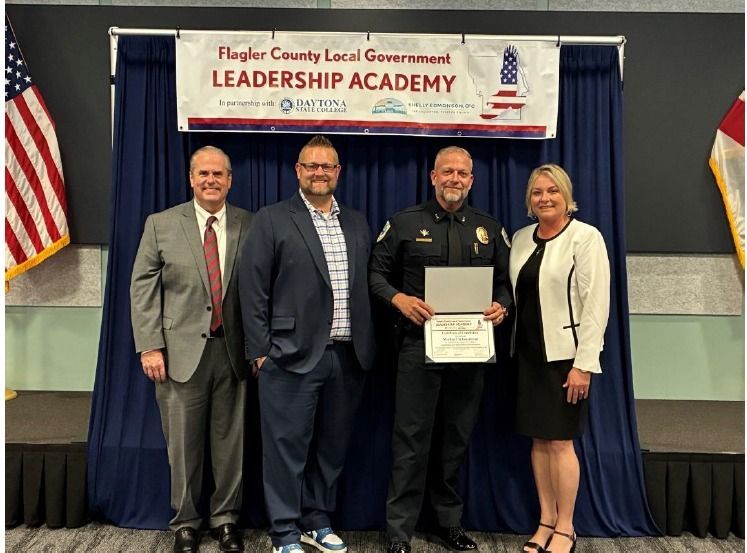 Four people at Flagler County Government Leadership Academy event, one holding a certificate on a stage