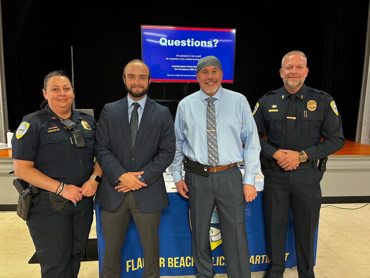 Four uniformed officers and a man in a suit stand smiling indoors at a briefing table.