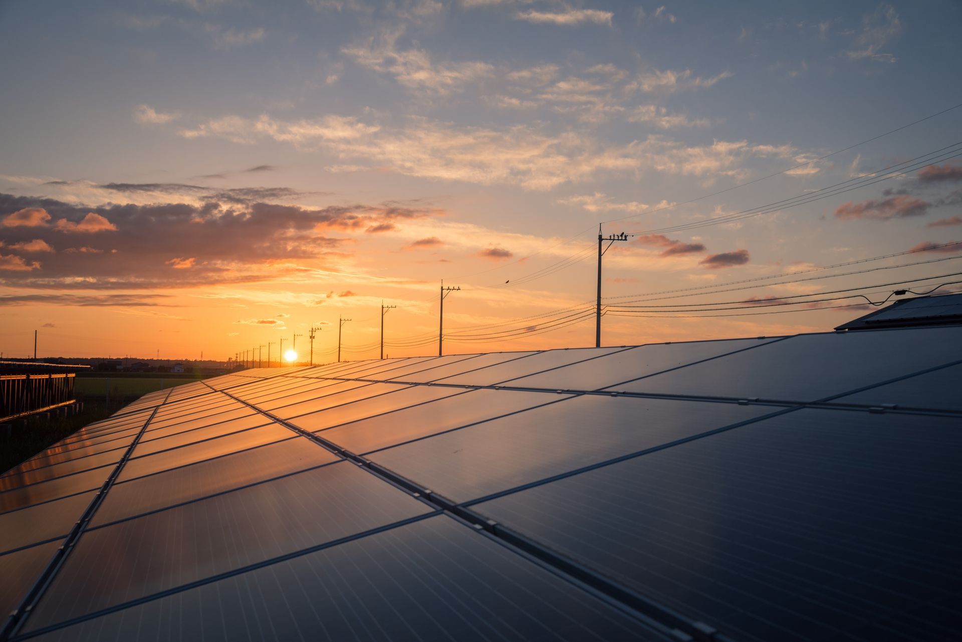 Solar panels in a field reflect the sunset, with utility poles and a colorful sky.