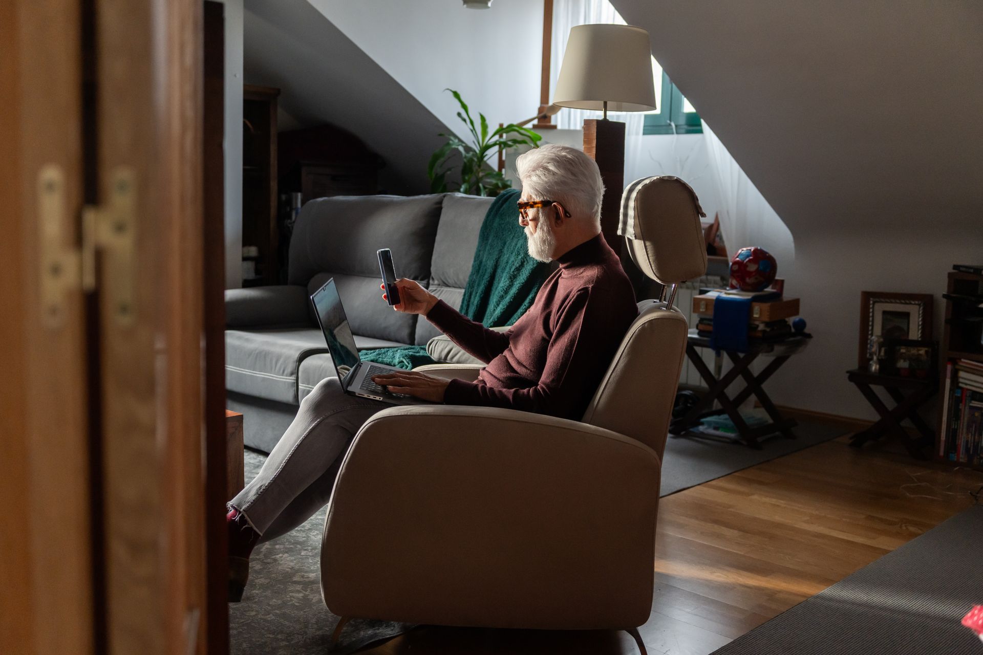 Elderly man seated in a chair, focused on using a laptop, with a thoughtful expression on his face.