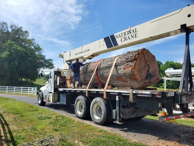 A crane truck on a rural road carries a large, massive cut log secured with straps, with an operator standing on the bed.