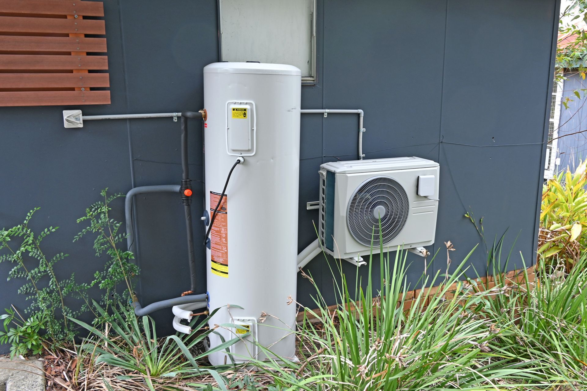 Outdoor utility equipment: white water heater and HVAC unit beside a dark gray building with plants in front — Hinch Plumbing In Breakwater, VIC