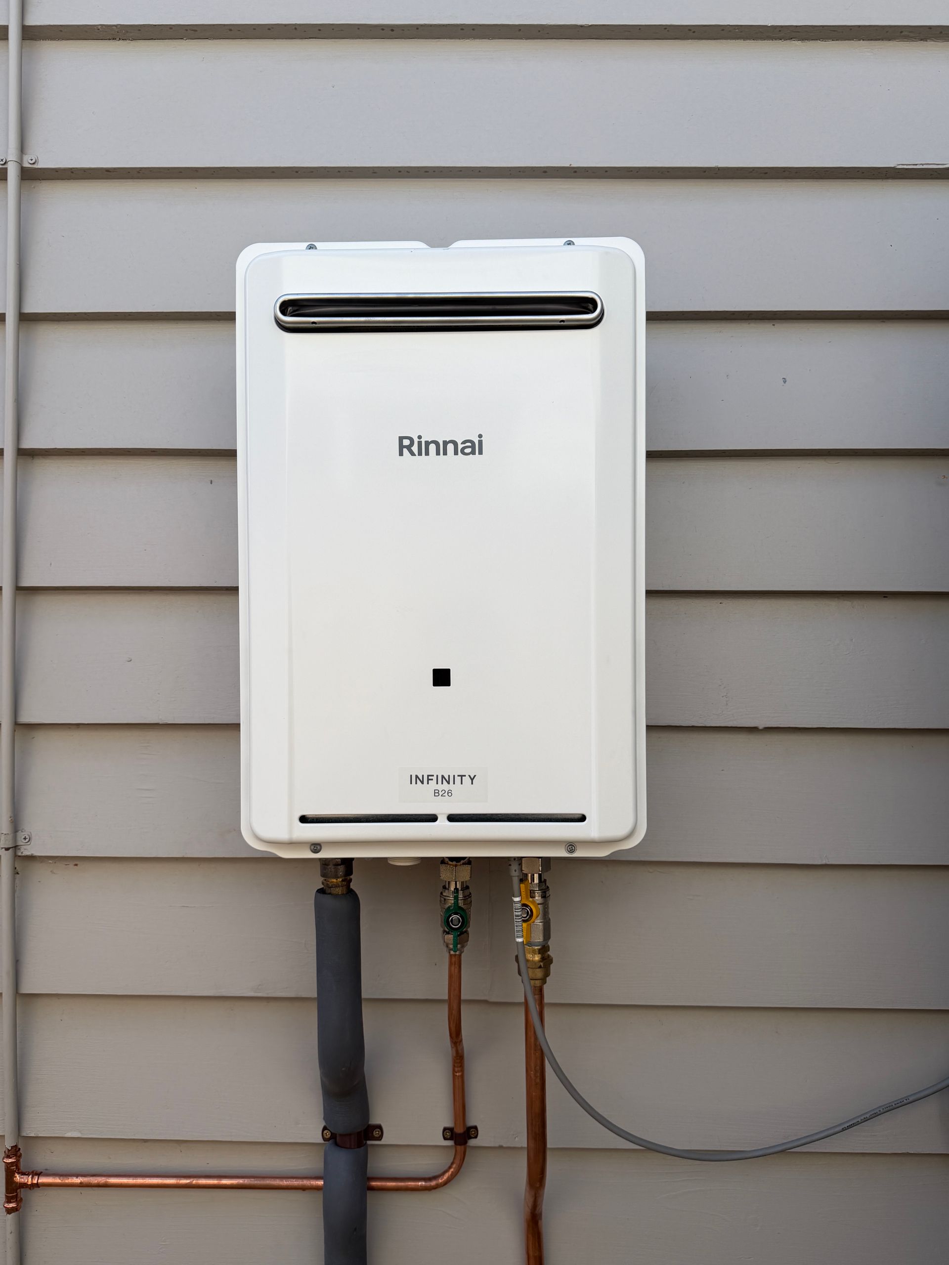 White Rinnai outdoor tankless water heater mounted on beige siding with connected pipes and hoses — Hinch Plumbing In Breakwater, VIC