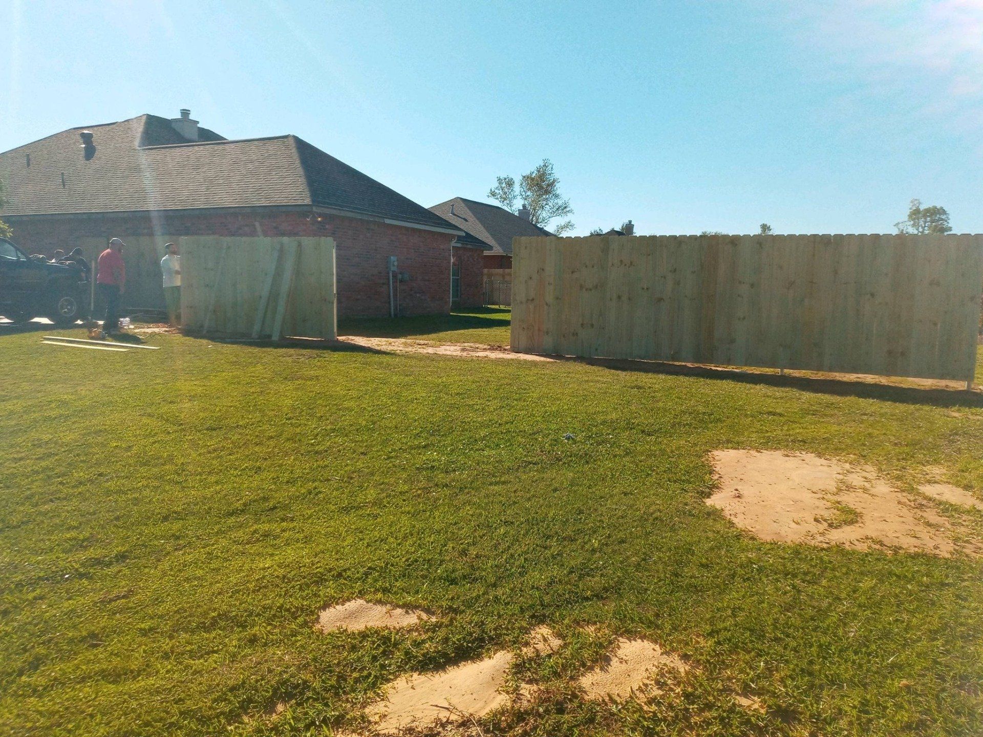 Backyard with green grass, tan fence, and brick house under a clear blue sky