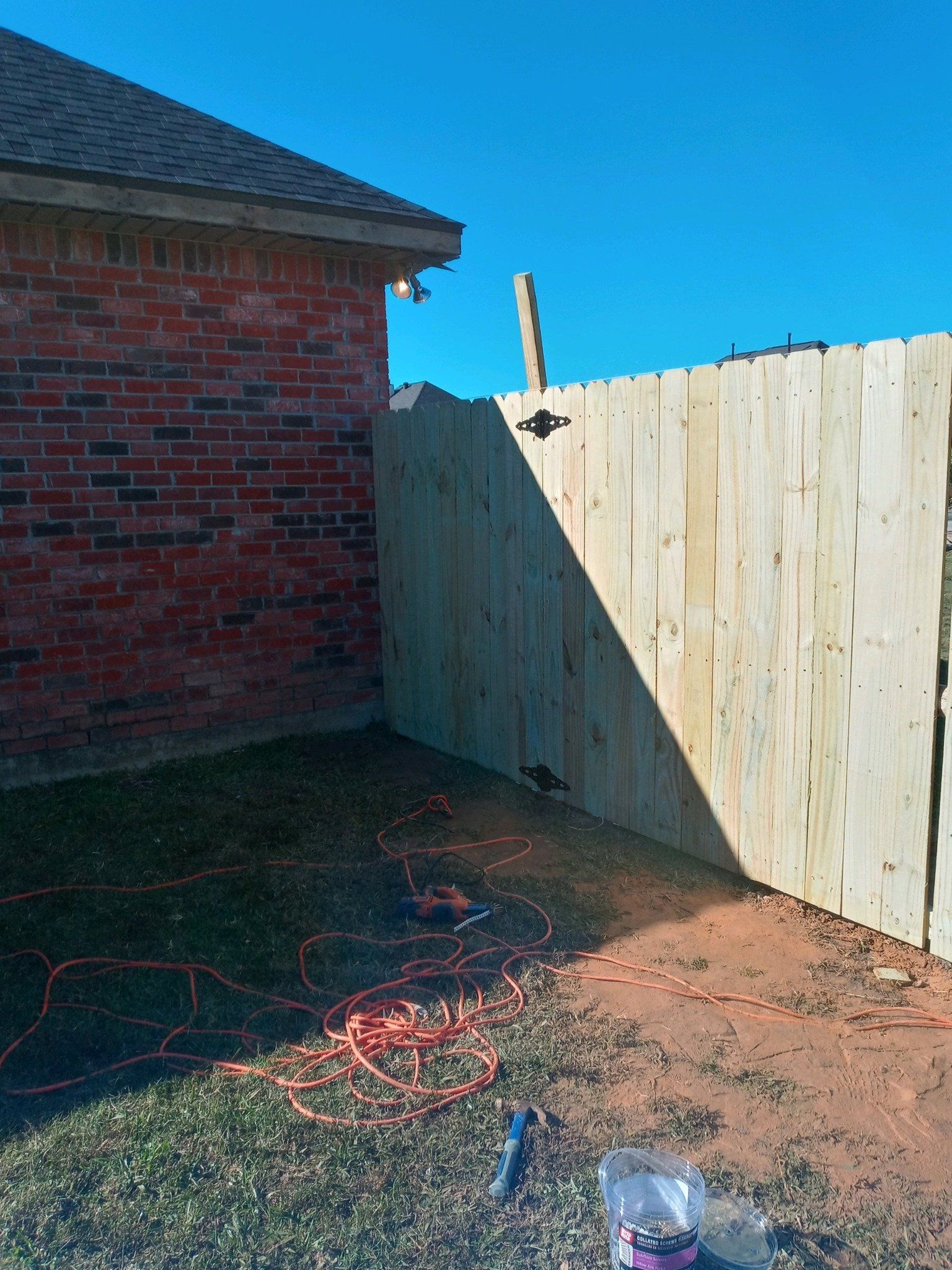 Backyard corner with brick wall, wooden fence, dirt ground, and scattered cables in sunlight