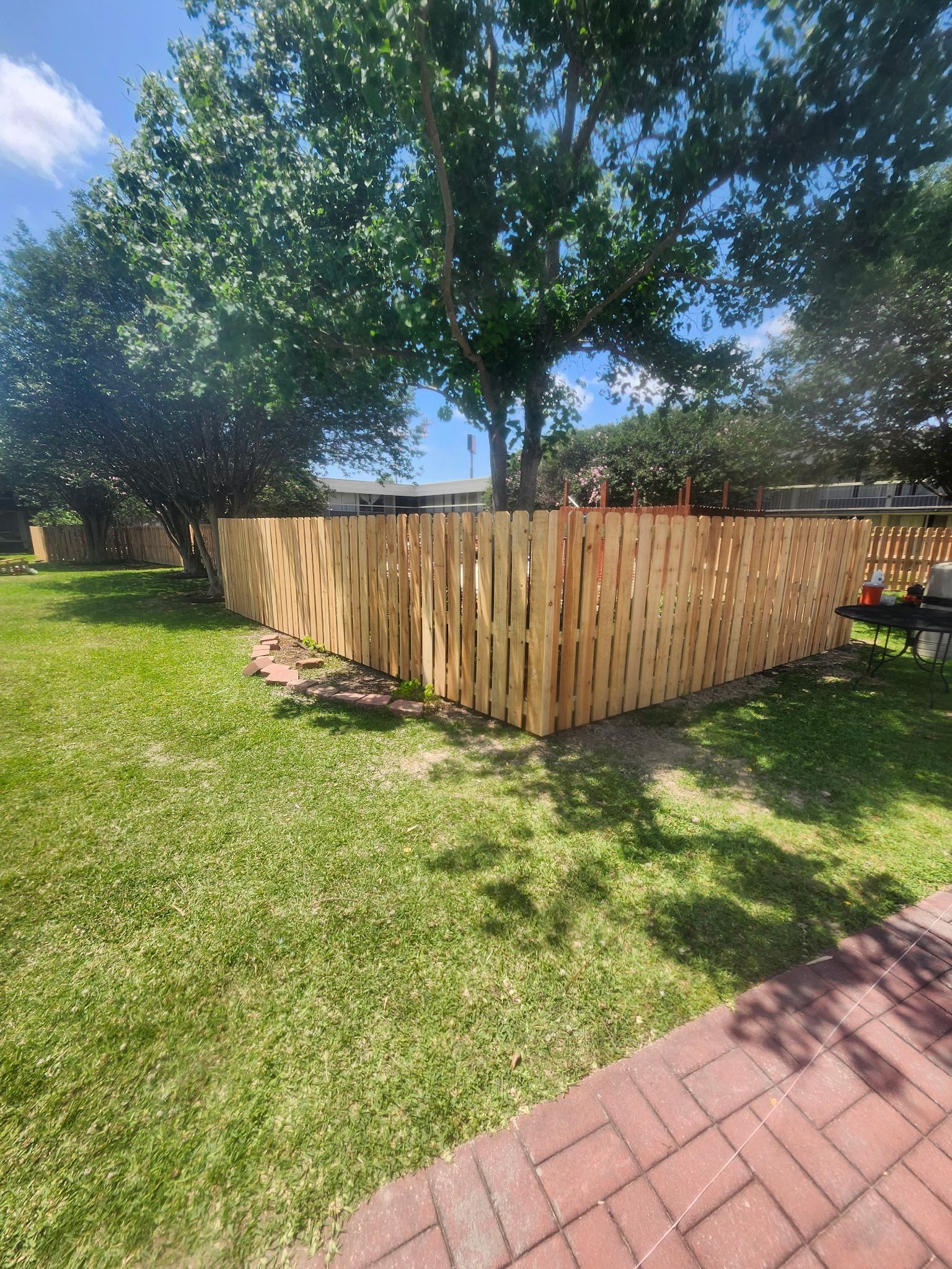 Wooden fence around a grassy yard with trees and a brick patio in bright sunlight