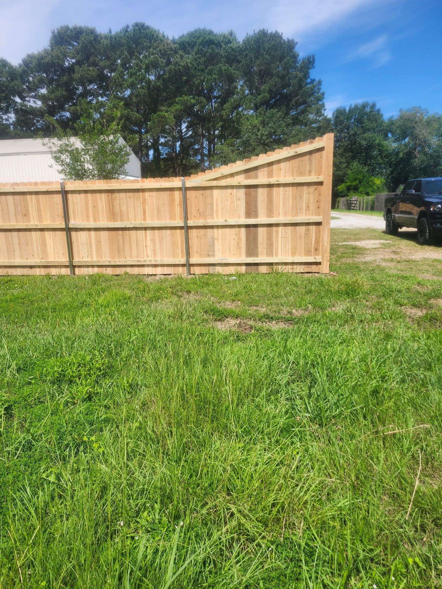 Wooden privacy fence beside a grassy yard under a blue sky, with a tree and parked truck in the background.