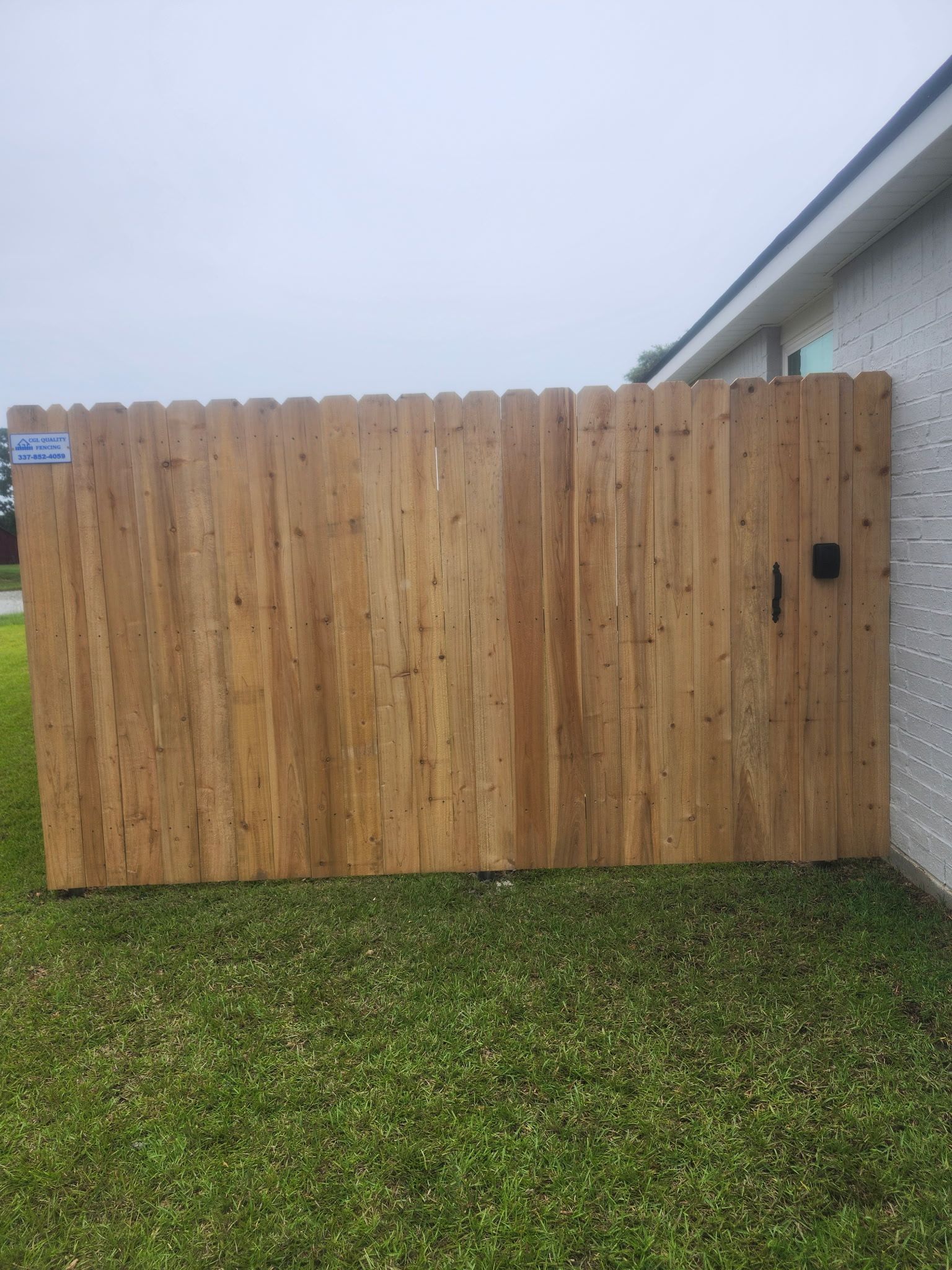 Wooden privacy fence beside a house, with green grass in the foreground and an overcast sky.
