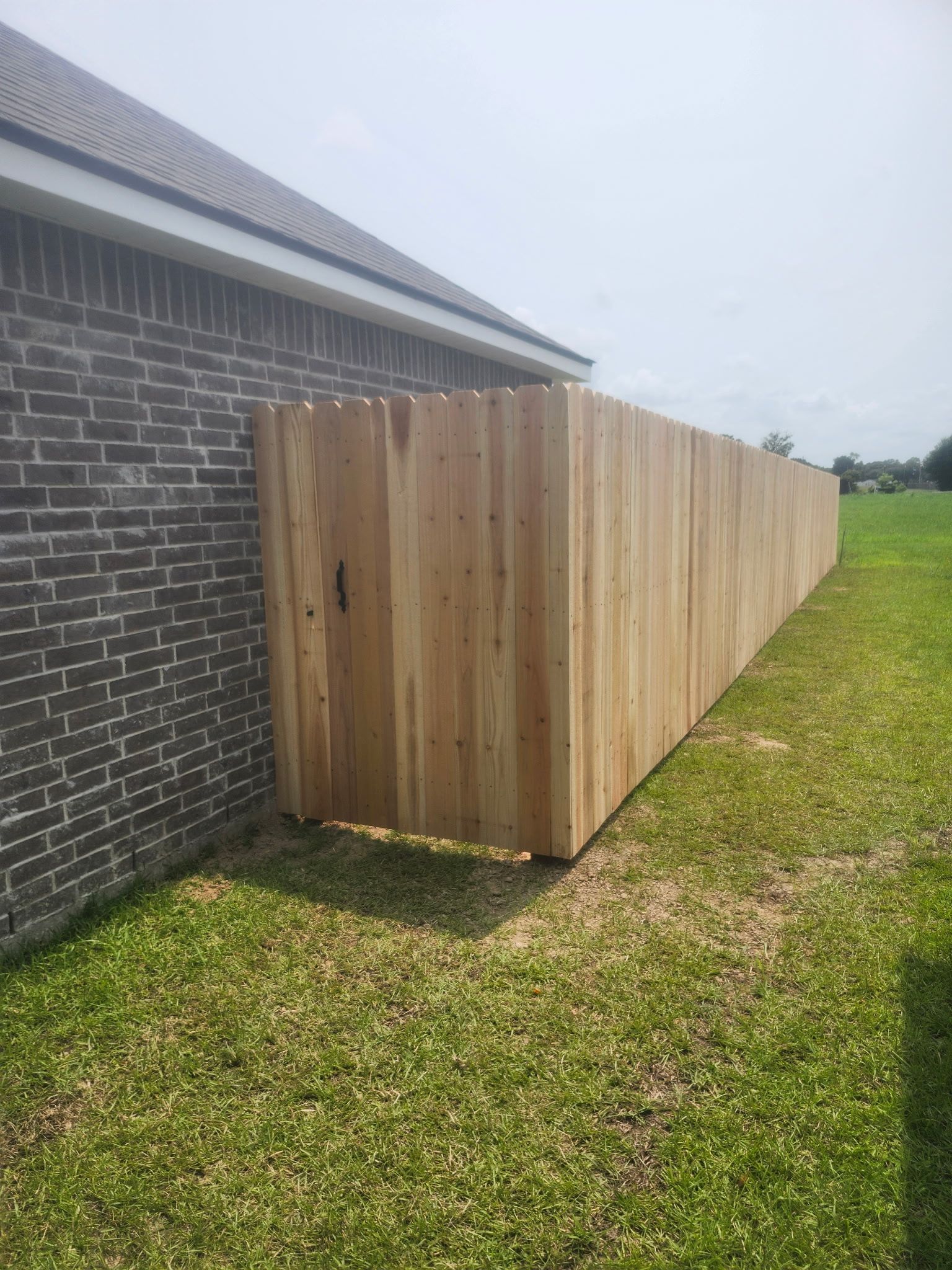 Long wooden privacy fence beside a brick house, running along a grassy yard under a gray sky