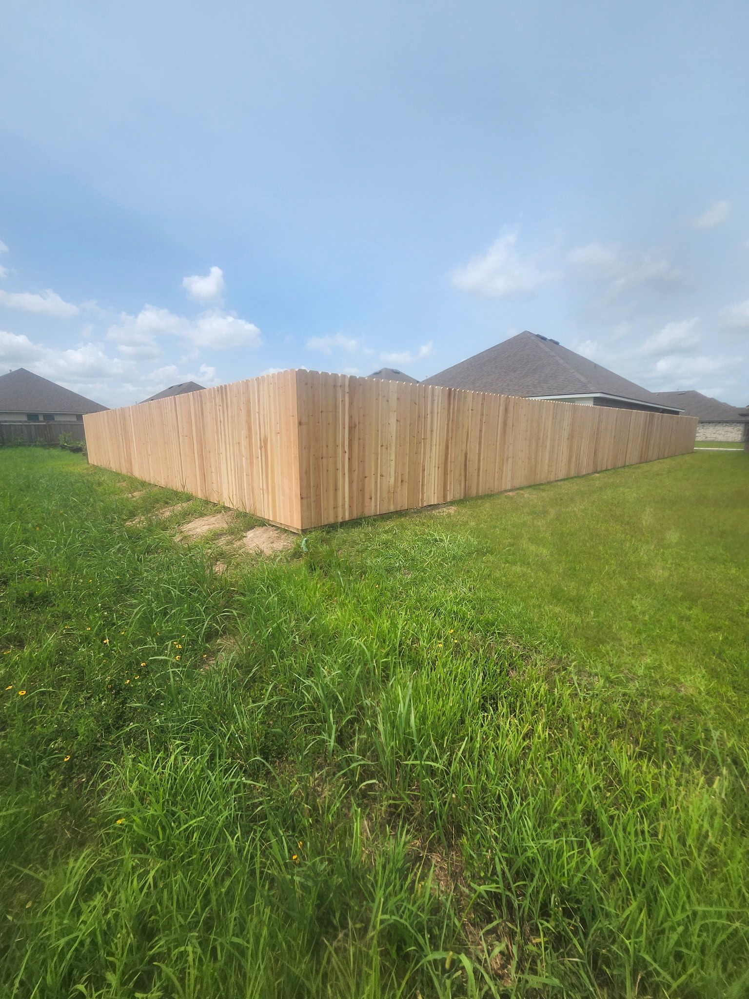 Wooden fenced enclosure in a grassy field under a blue sky, with rooftops in the background
