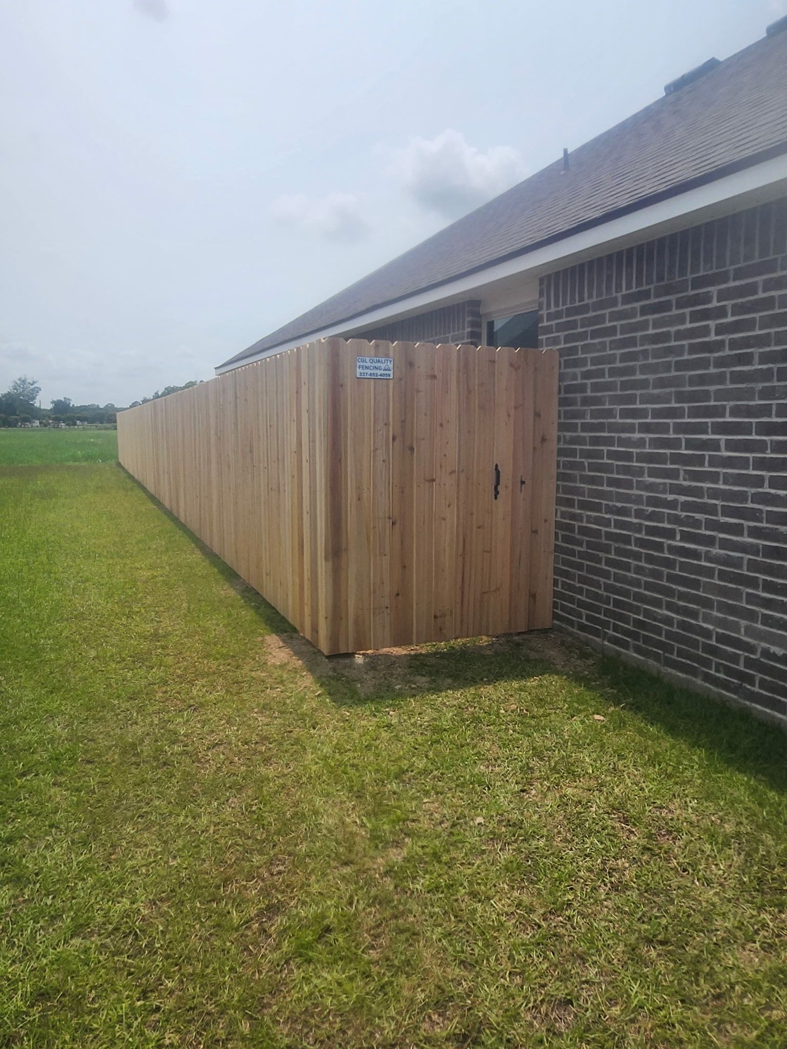Wooden privacy fence beside a brick house, extending across a grassy yard under a cloudy sky
