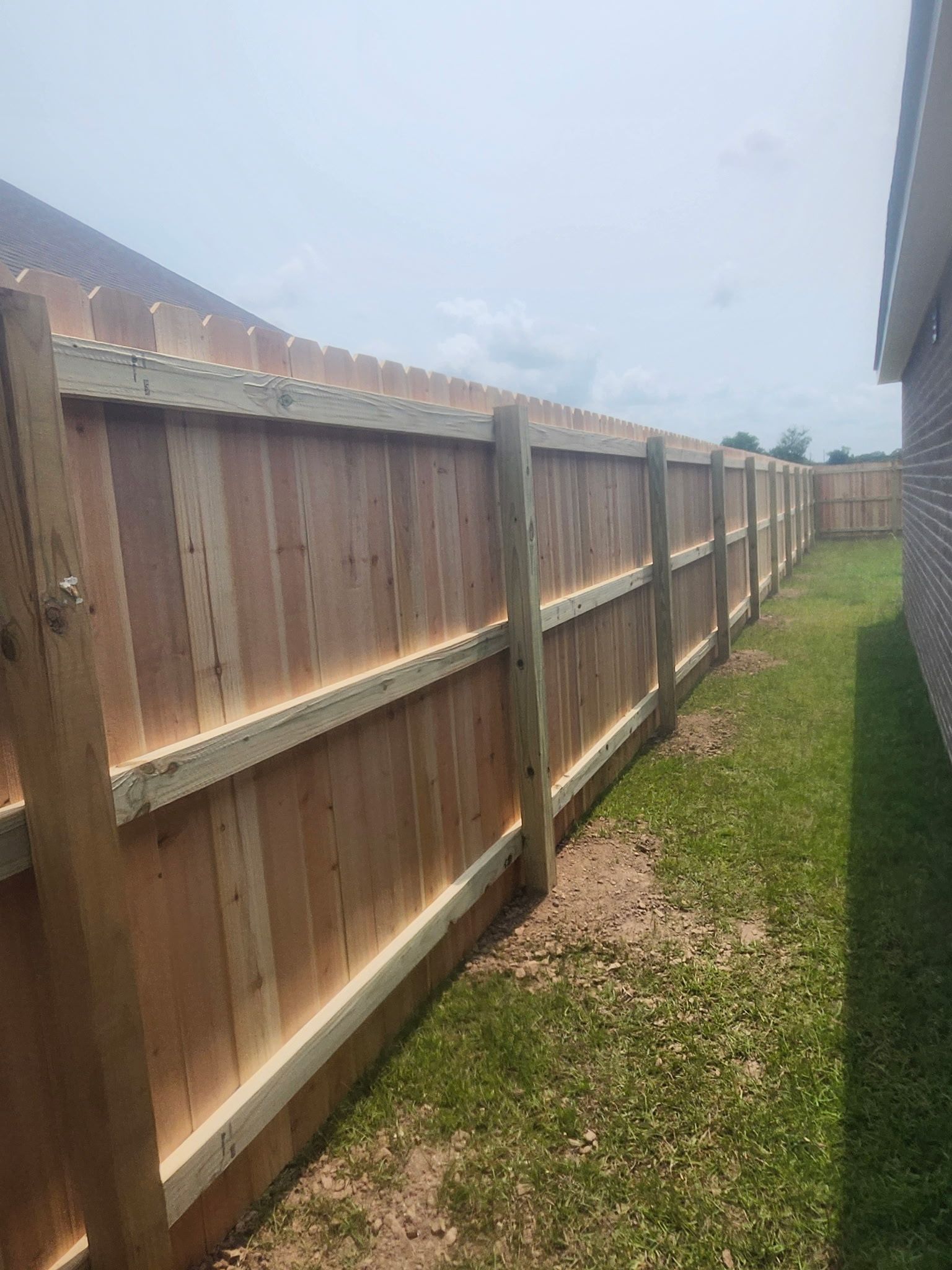 Wooden backyard fence along a narrow grassy side yard beside a house under a bright sky