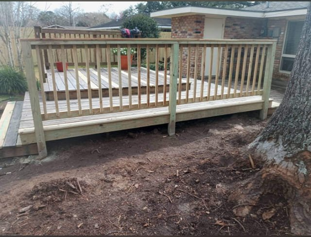 Wooden accessibility ramp with railings beside a house, over bare dirt near a large tree.