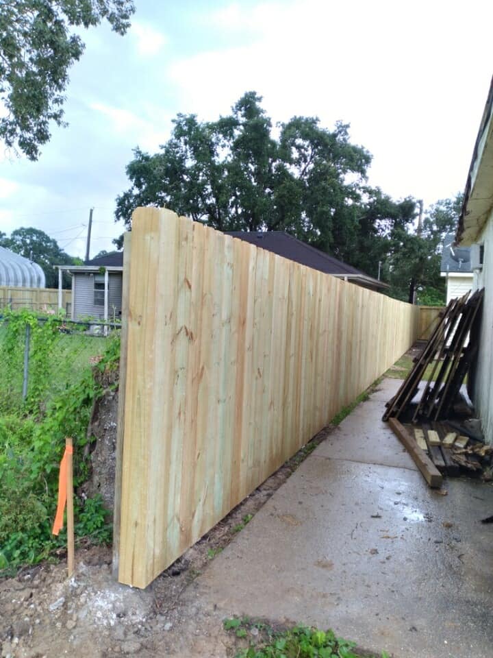Long wooden fence along a wet concrete path beside stacked lumber and greenery