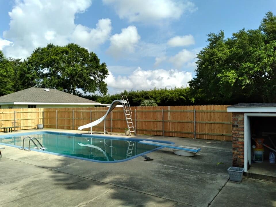 Backyard pool with a slide, wooden fence, and open shed under a partly cloudy sky