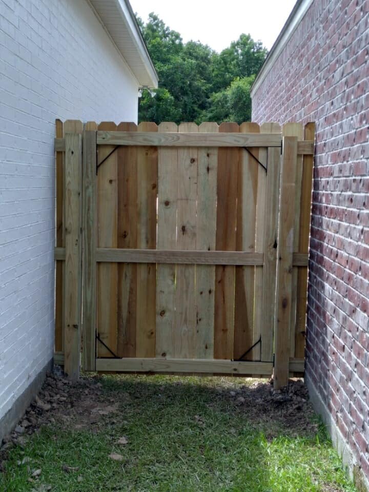 Wooden gate blocking a narrow grassy passage between a white wall and a brick wall.