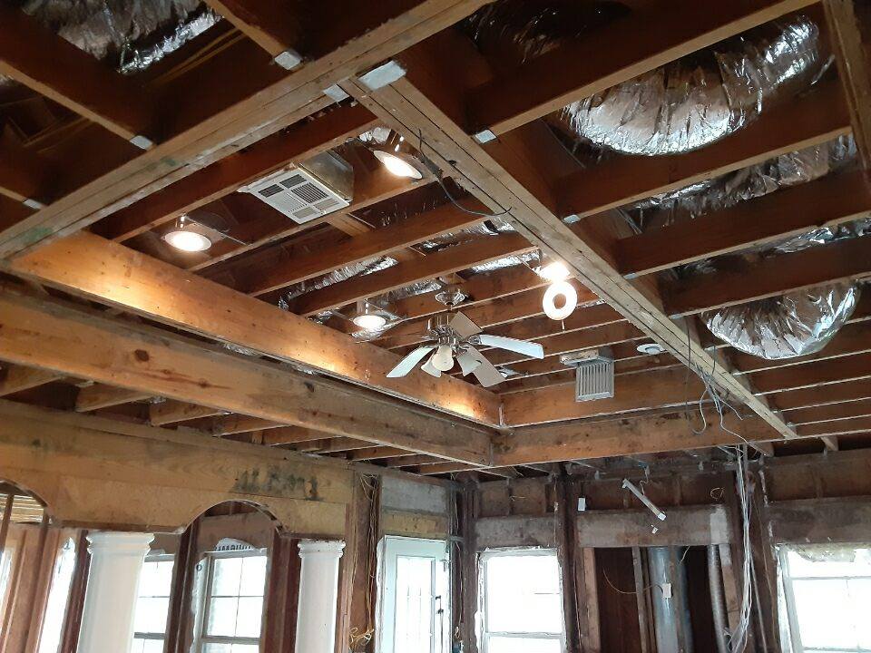 Damaged room ceiling with exposed wooden beams, insulation, and hanging light fixtures above windows