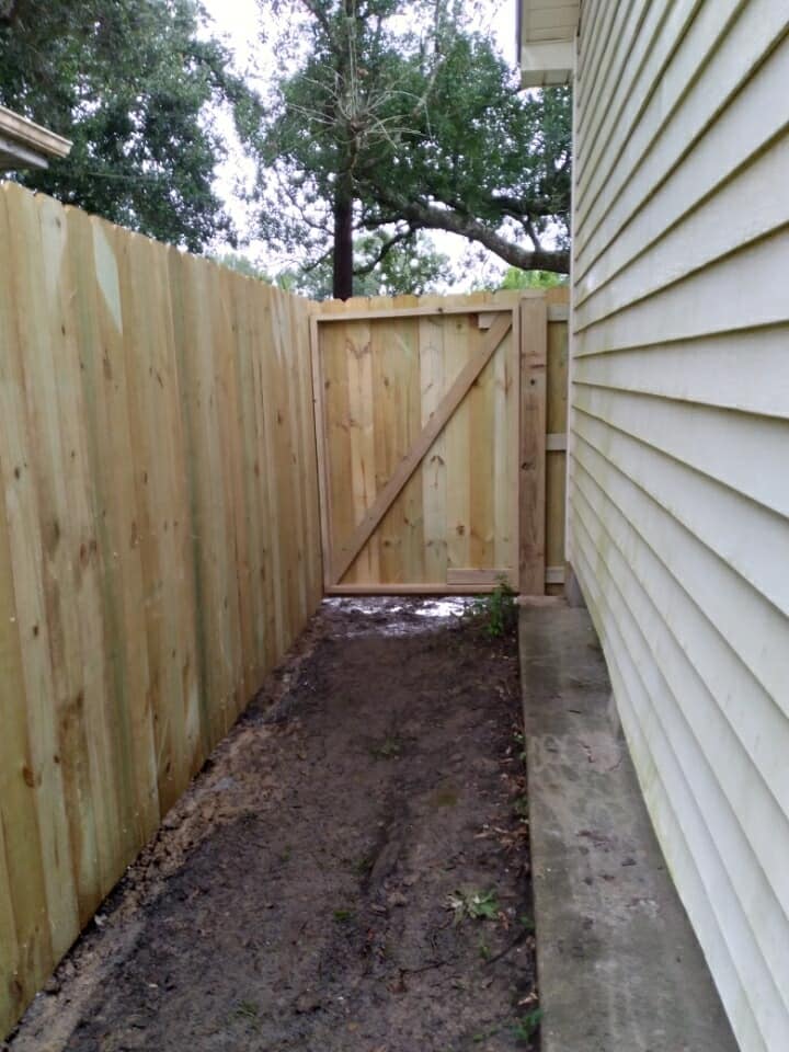 Narrow dirt path beside white house and wooden fence leading to a closed gate.