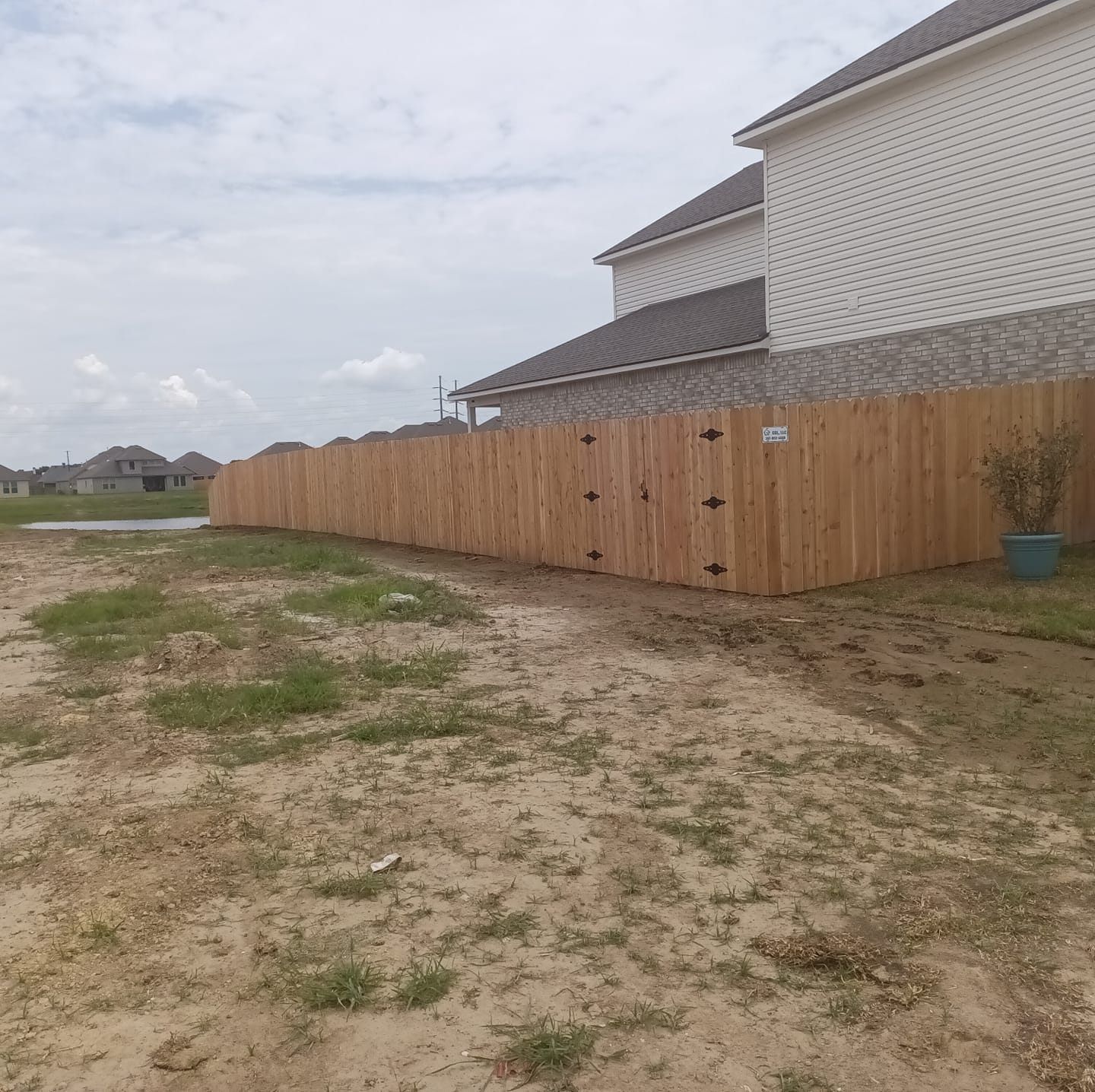 Wooden privacy fence beside a white house on a sandy vacant lot under a cloudy sky