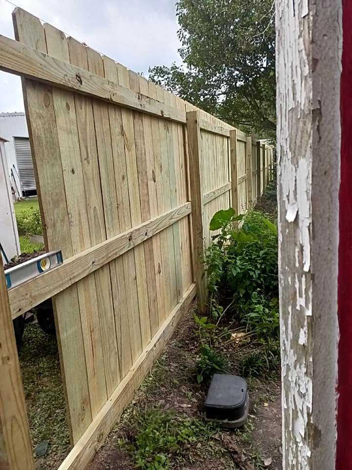 Narrow side yard with a new wooden fence, bushes, and a small black box on the ground