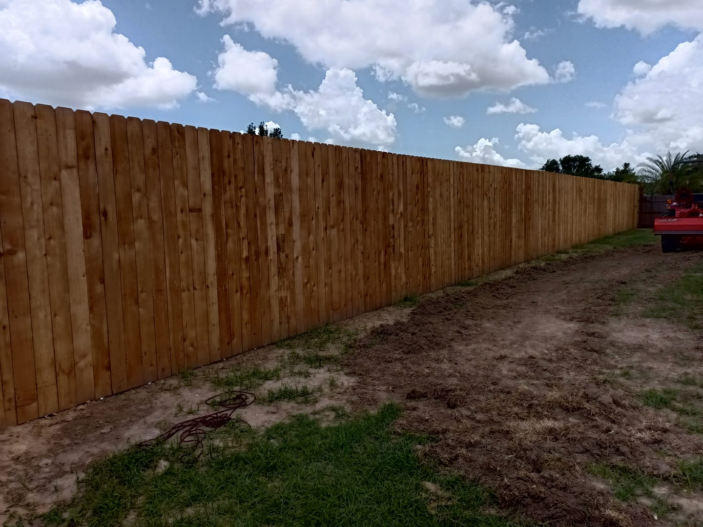 Long wooden privacy fence along a patchy dirt yard under a partly cloudy sky.