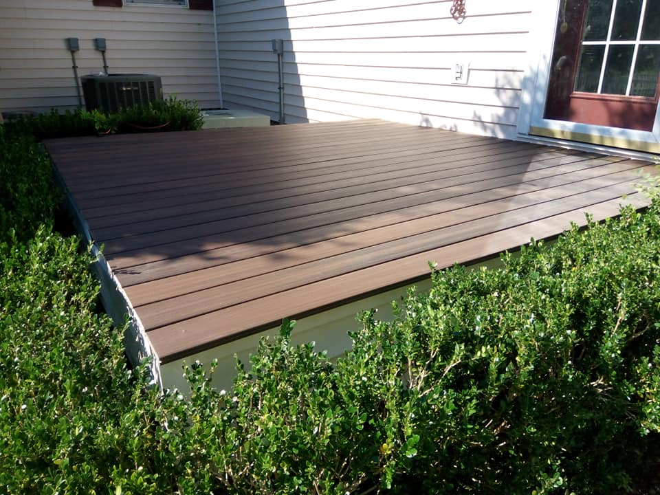 Wooden backyard deck beside a house, bordered by green shrubs in sunlight.
