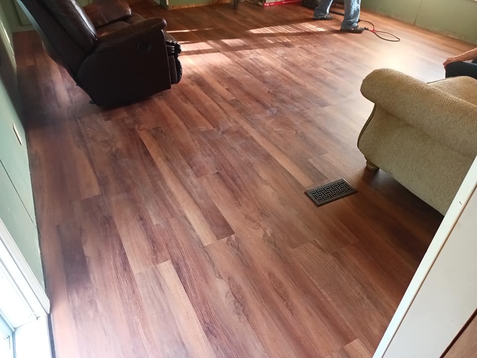 Living room with wood flooring, a dark sofa, beige chair, and sunlight on the floor.