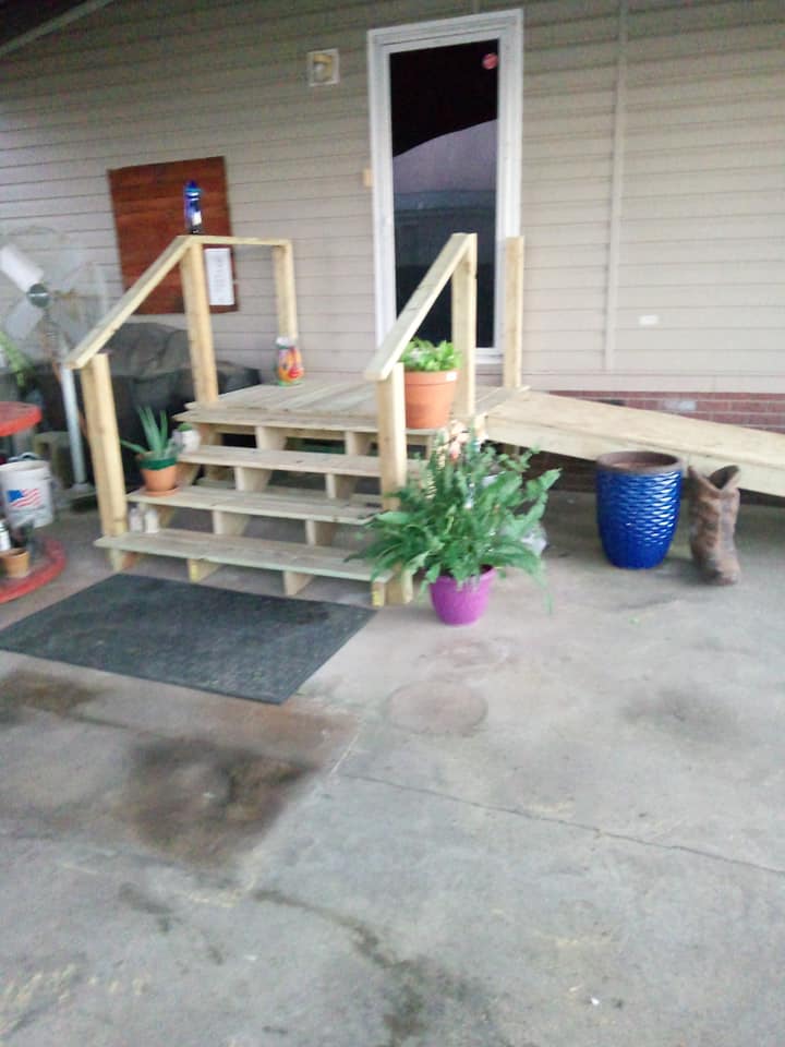 Front porch with wooden steps, handrails, potted plants, a blue planter, and a cat on the right side.