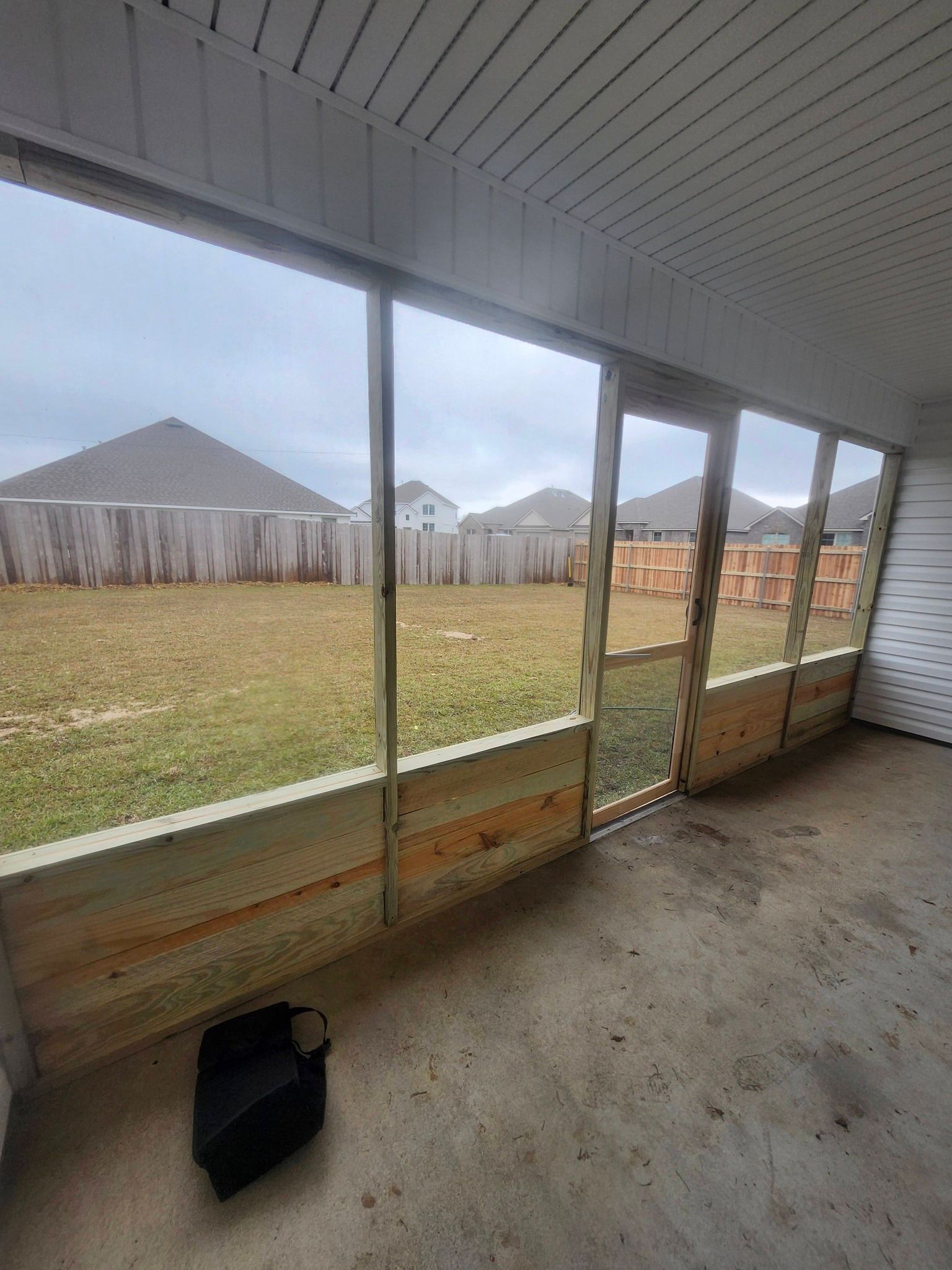 Covered patio with large windows overlooking a fenced backyard and neighboring houses on a cloudy day