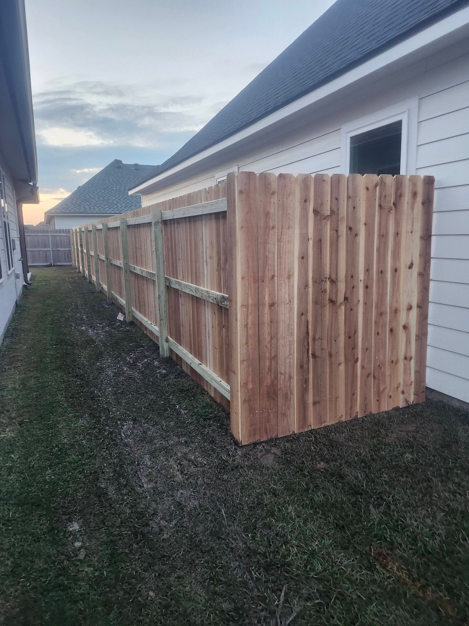 Wooden privacy fence along a narrow side yard beside a house under a cloudy sky.