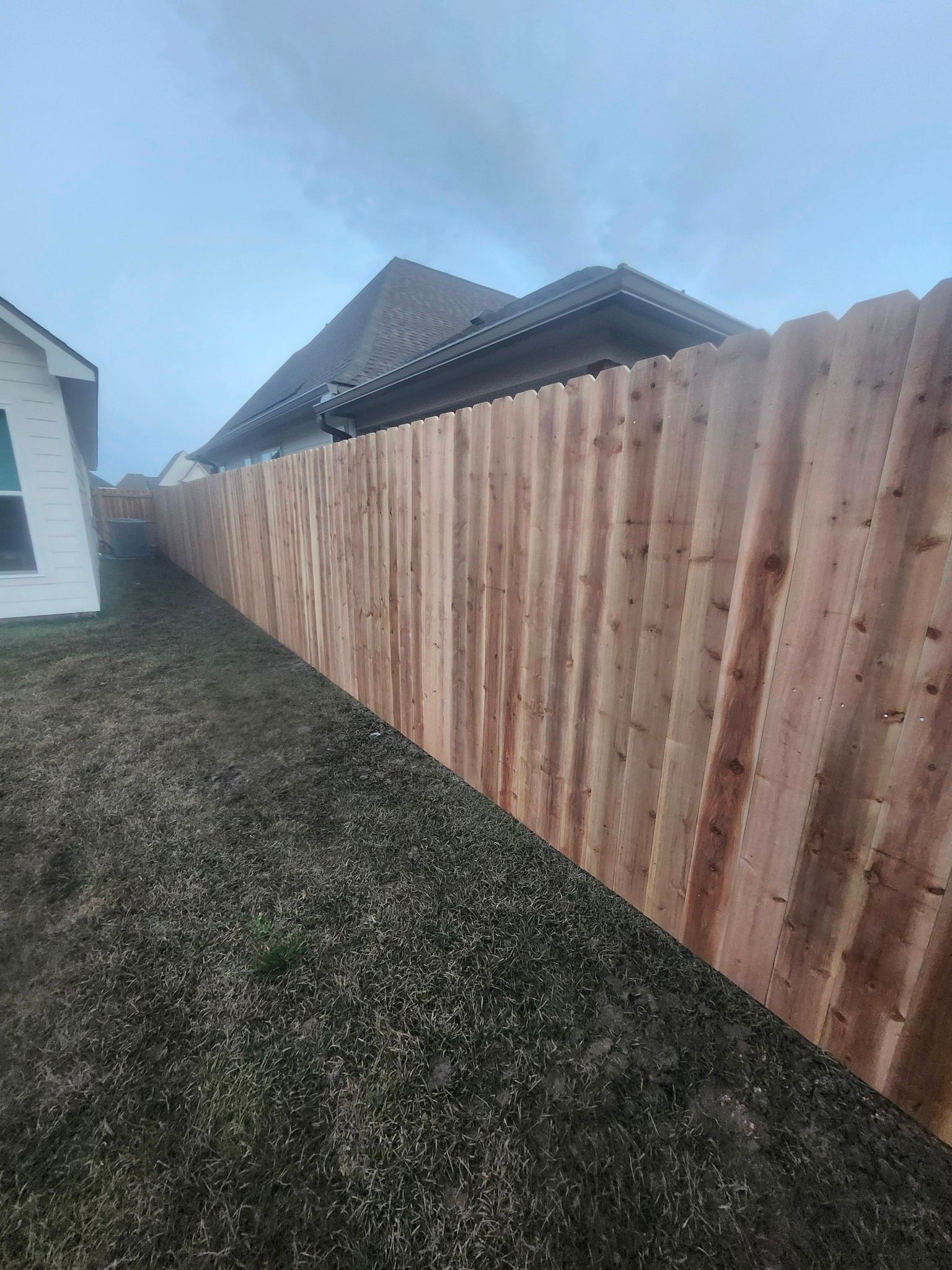 Long wooden fence beside a gravel path, with house roofs under a cloudy sky.