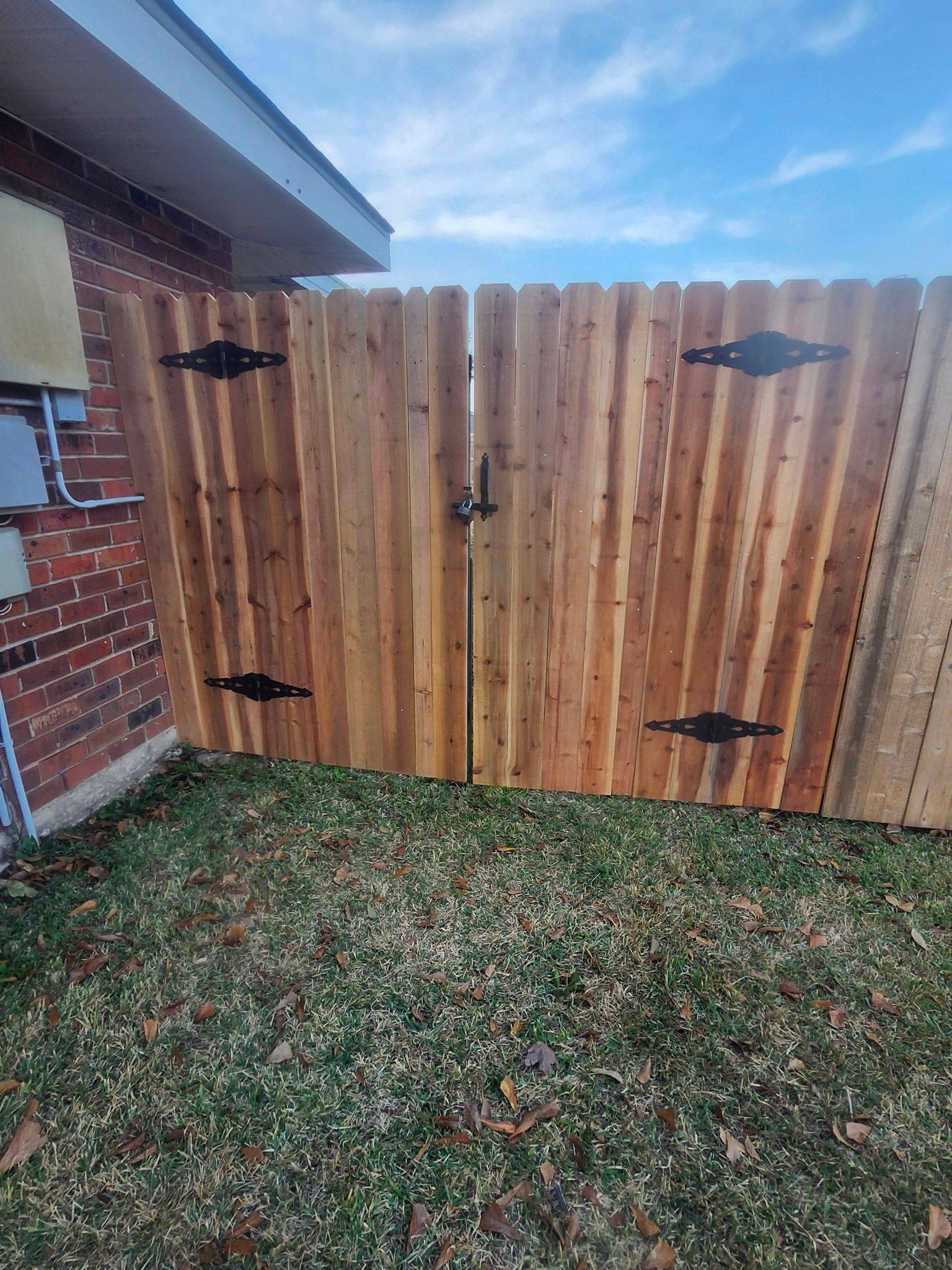Wooden backyard fence with black hinges beside a brick house and patchy grass lawn
