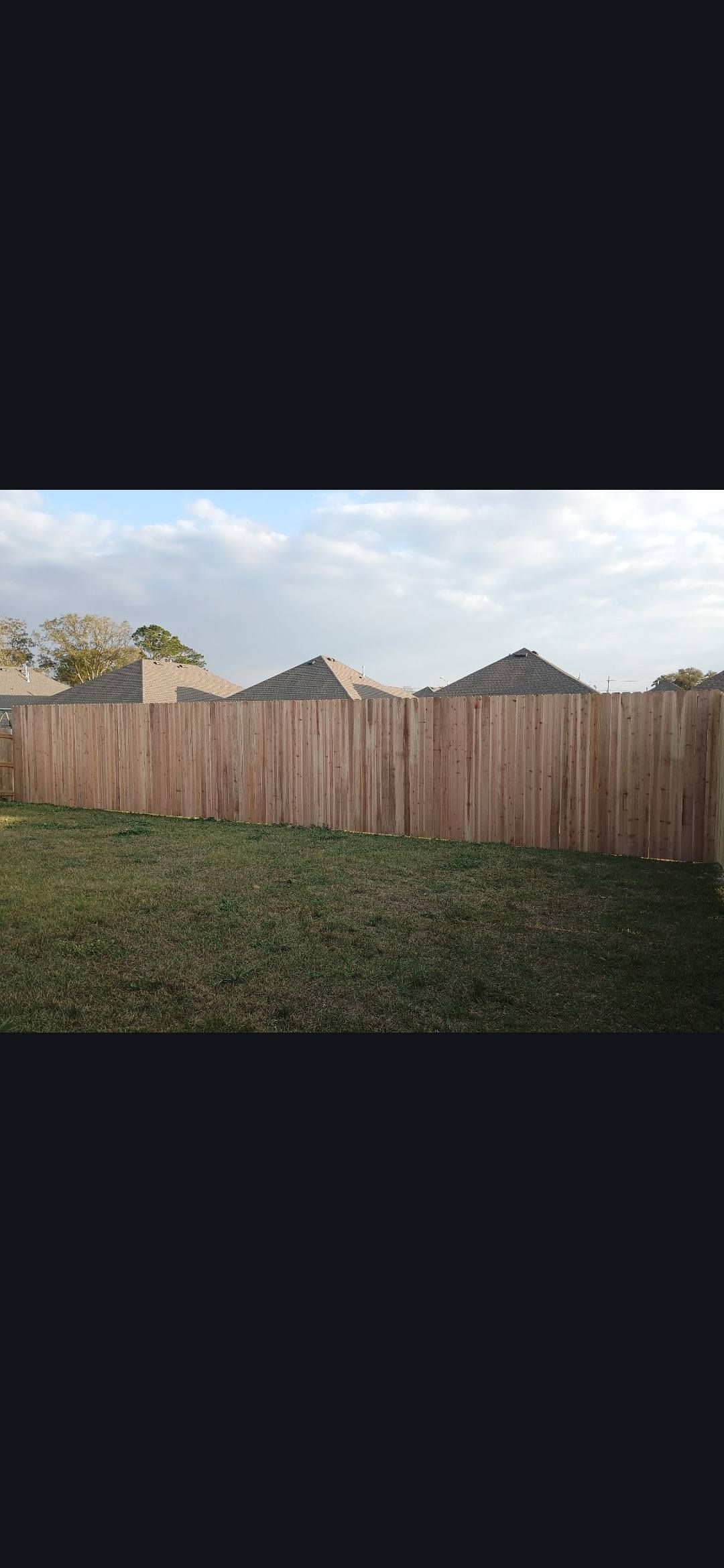 Backyard with a tall wooden fence, grassy lawn, and rooftops under a cloudy sky