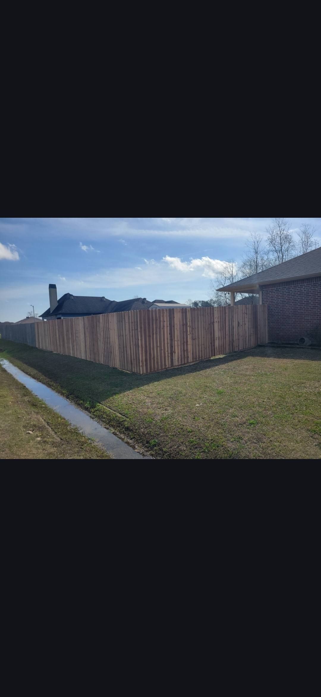 Residential sidewalk beside a tall wooden fence under a bright blue sky with clouds