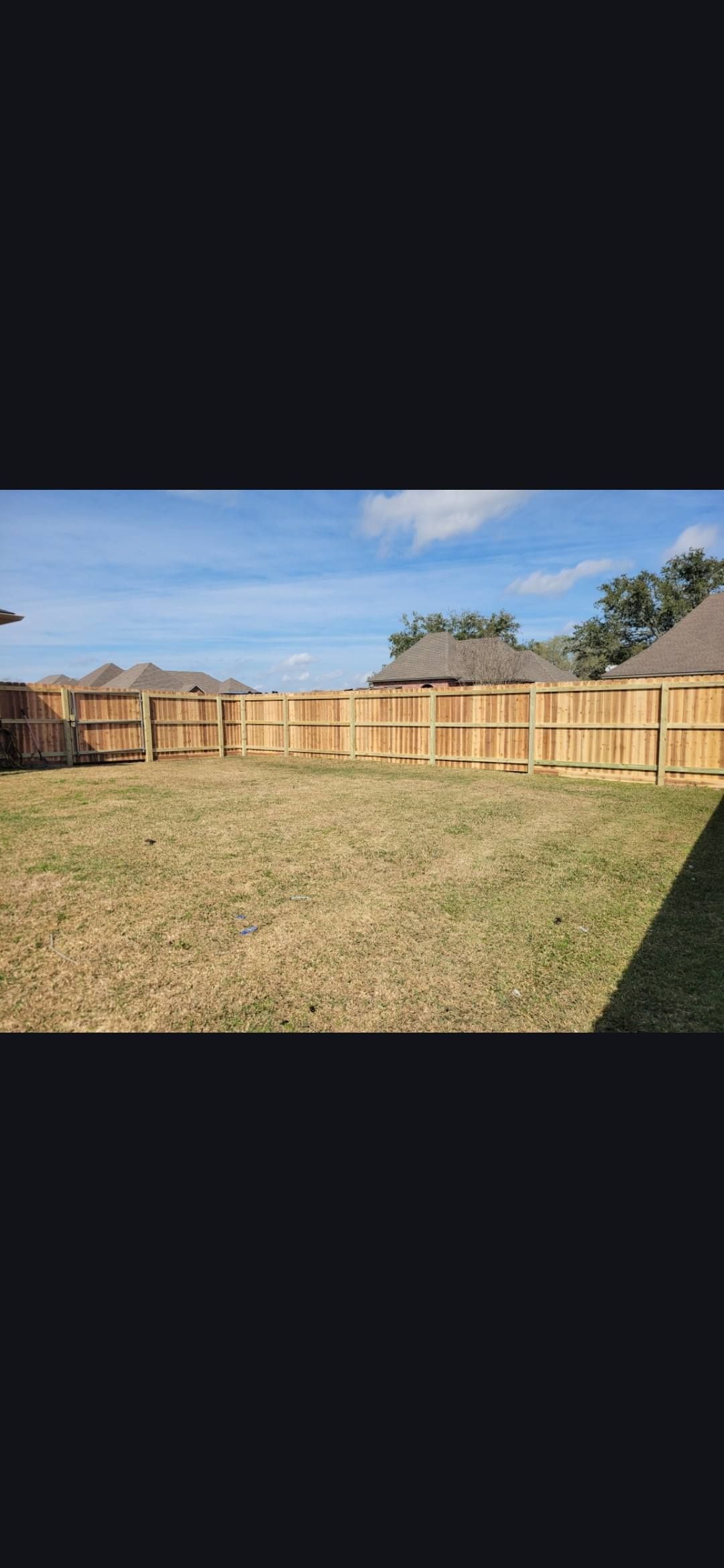 A fenced grassy backyard under a blue sky with trees and neighboring houses in the distance.