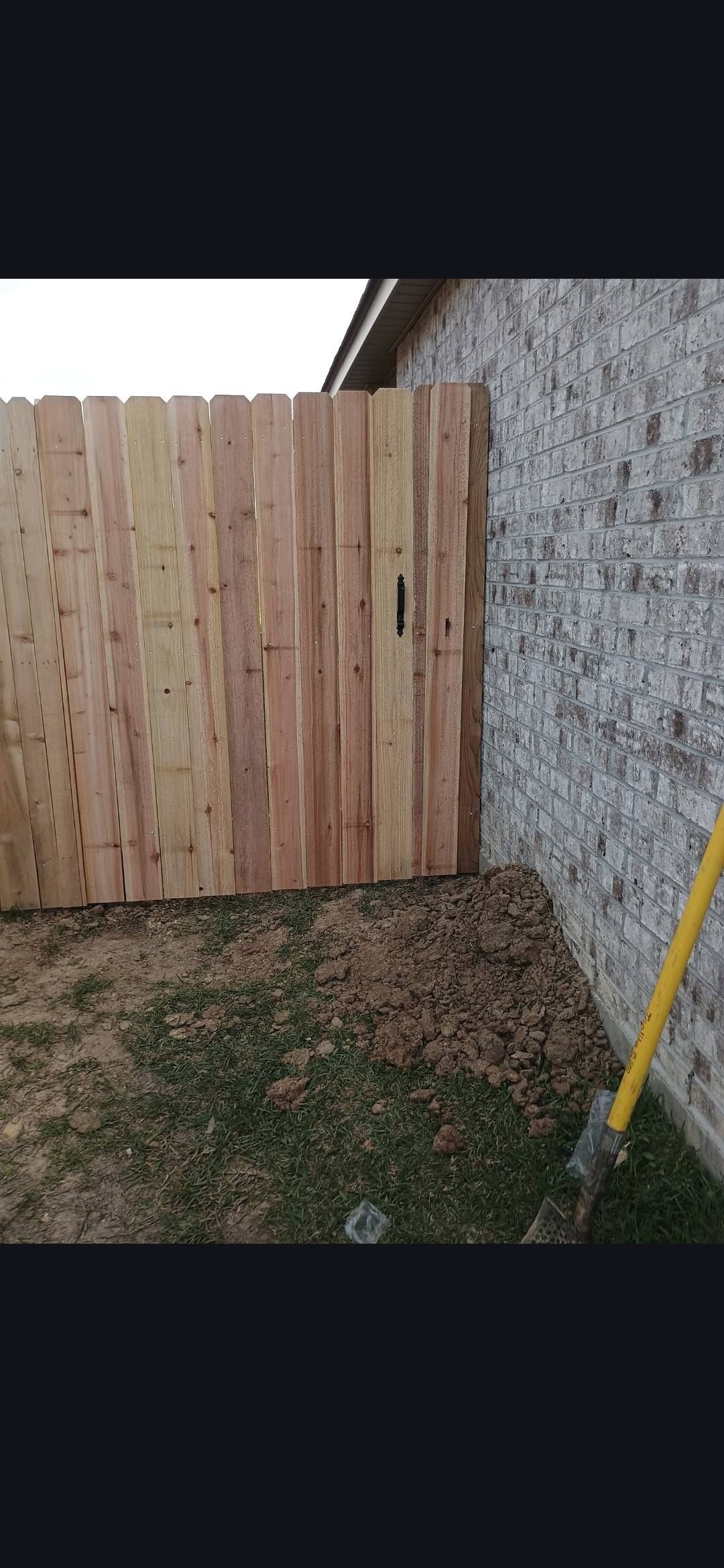 Wooden fence beside a gray wall and patchy grass, with a yellow-handled tool at the right edge.