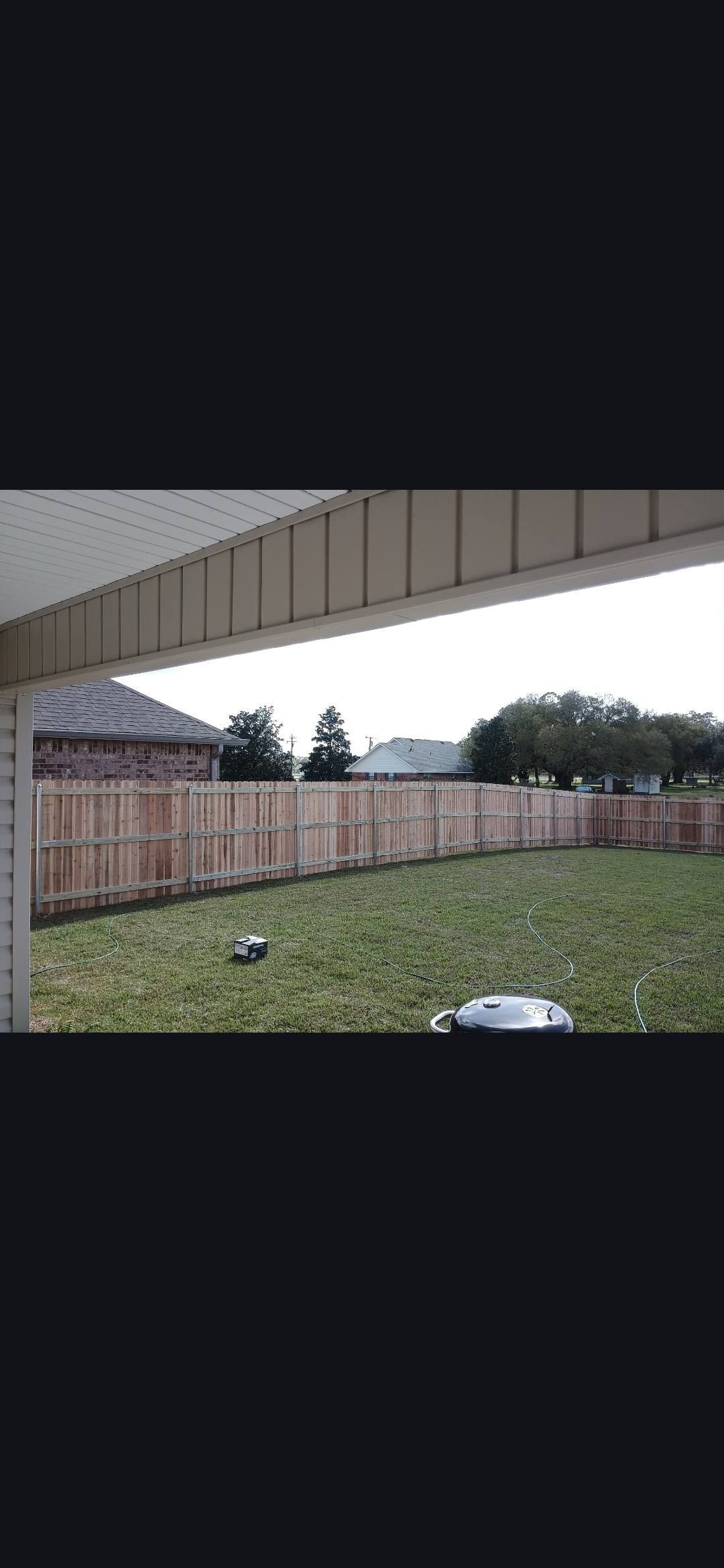 Backyard under a covered patio, with a wooden fence, grass, and overcast sky.