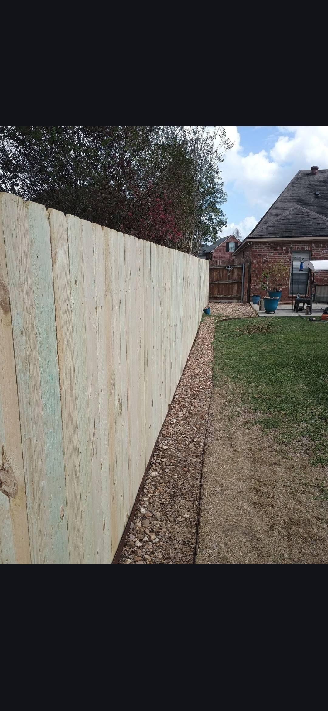 Wooden fence beside a dirt path and grassy yard, with trees and a house in the background.