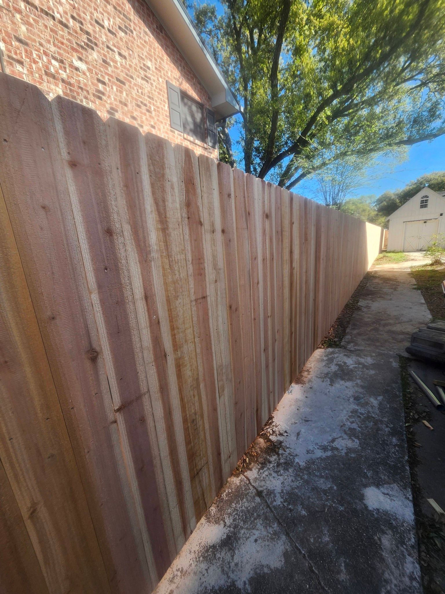 Narrow side yard with a tall weathered wooden fence beside a brick wall under a blue sky.