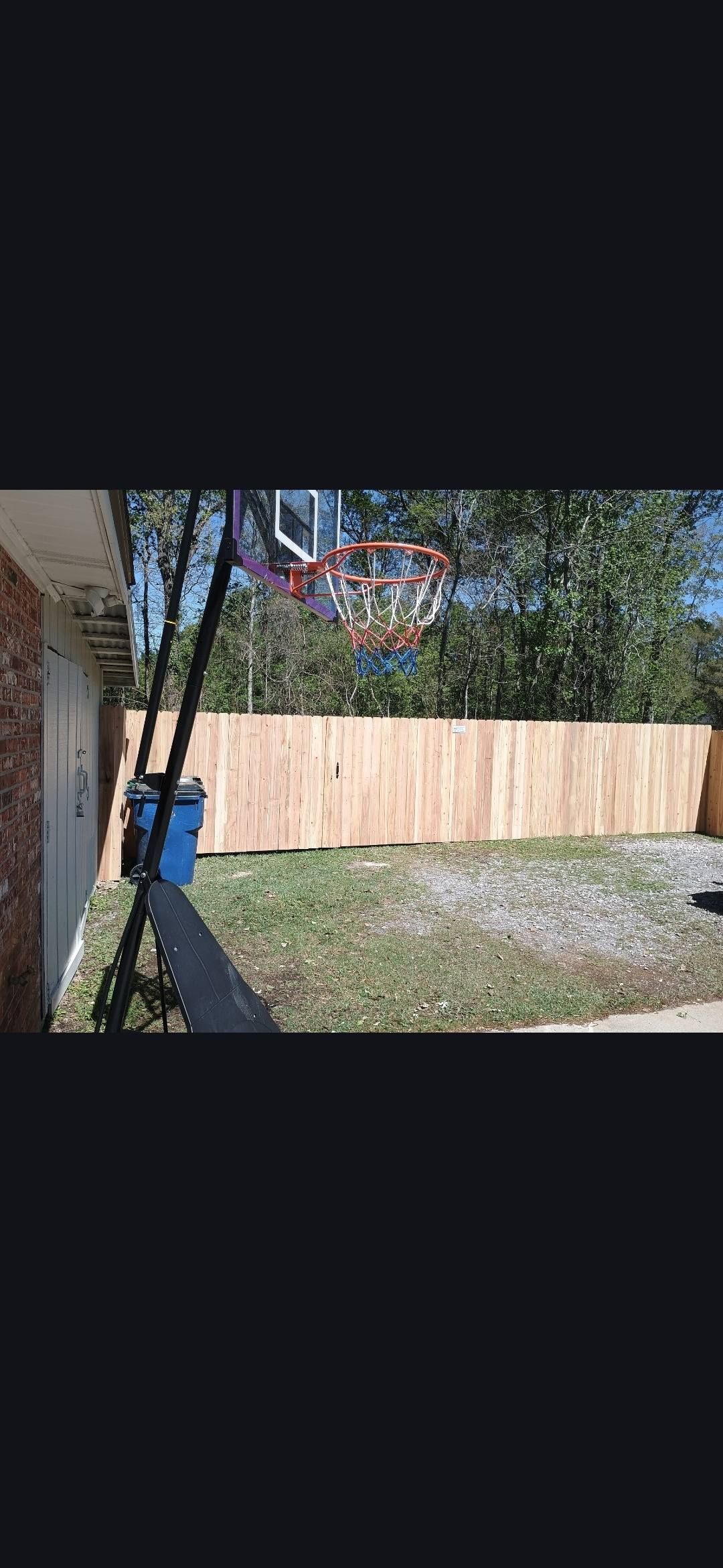 Backyard with a wooden fence, trees, gravel ground, and a blue trash bin beside a house.